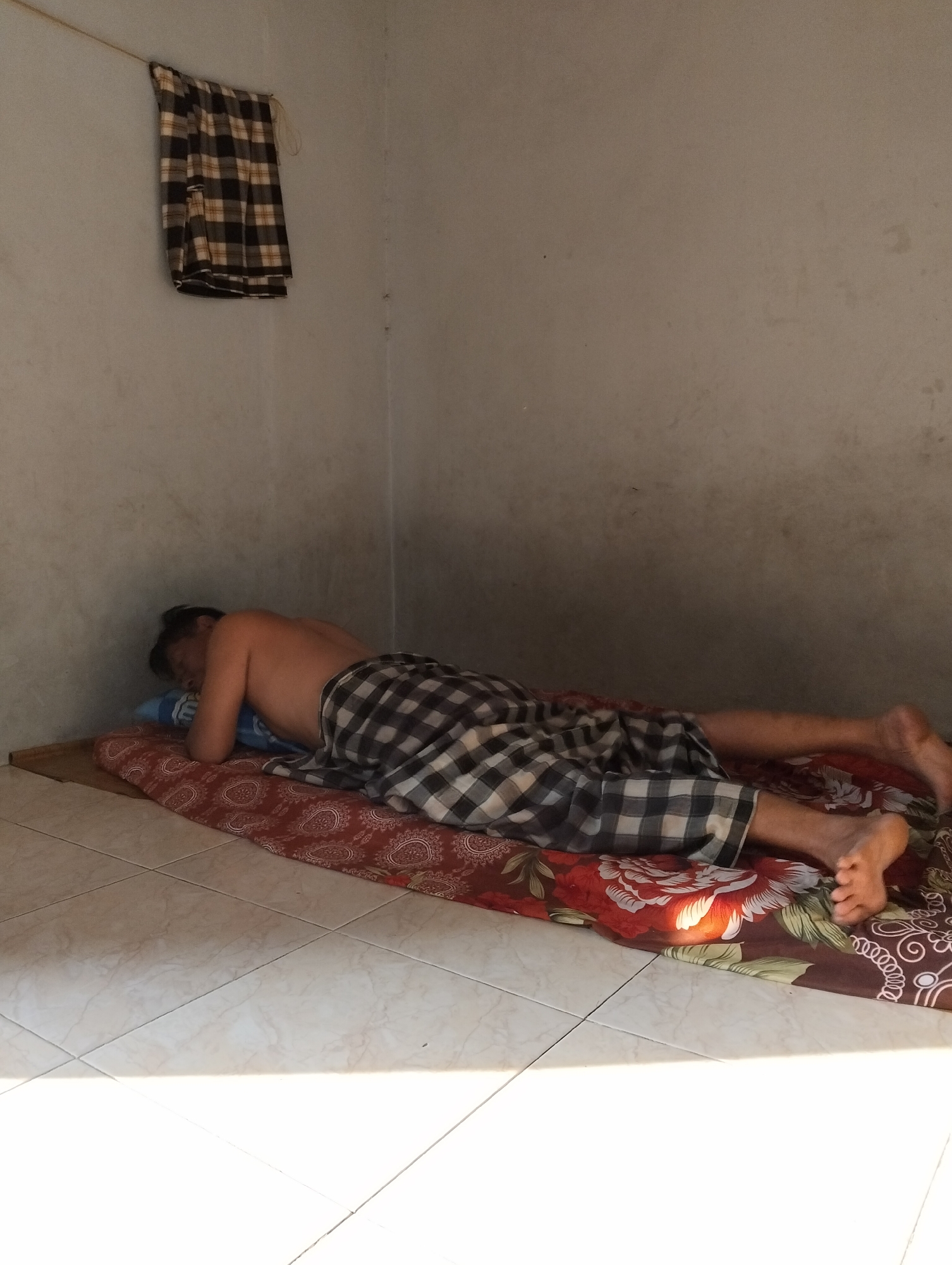 A person is lying face down on a bed in a sparsely furnished room. The person is wearing checkered pants and appears to be sleeping. The bed is covered with a patterned sheet and a thin mattress. A towel with a checkered pattern is hanging on the wall above the bed. The room has white walls and a tiled floor. A shadow from a window is cast across the floor. The scene is likely in a residential setting in Gambut, Indonesia, given the style of the room and the common practice of drying laundry indoors or in covered areas.