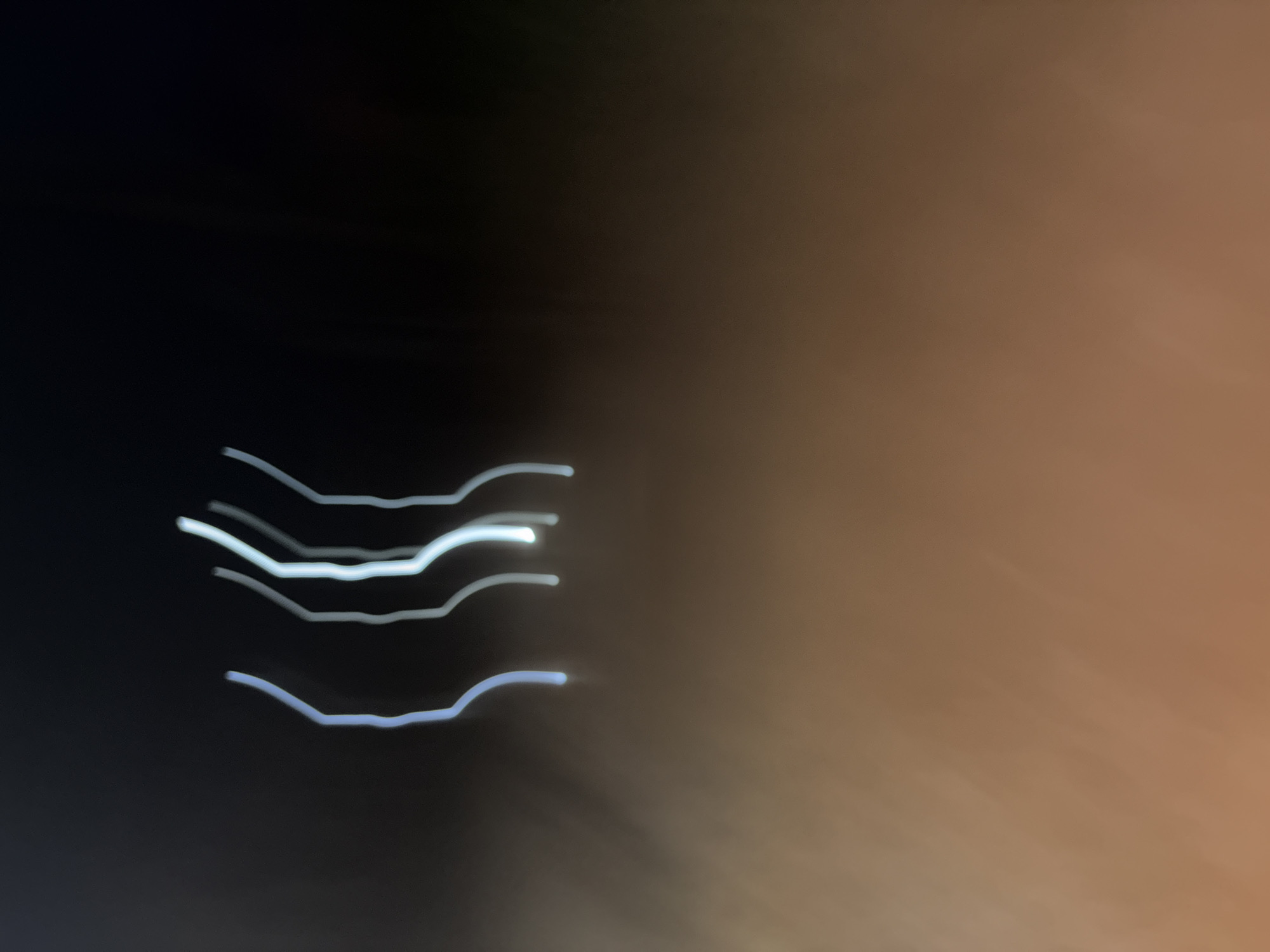 The image is an abstract, long-exposure photograph taken at night in Obosi, Nigeria. It features several blurred, glowing white lines against a dark background that transitions to a warm, brown hue at the bottom. The lines are vertical and appear to be light trails, possibly from moving vehicles or other light sources. There are no discernible people, objects, or specific landmarks visible in the image due to the motion blur. The environment appears to be an outdoor urban setting at night, characterized by artificial lights and a dark sky.