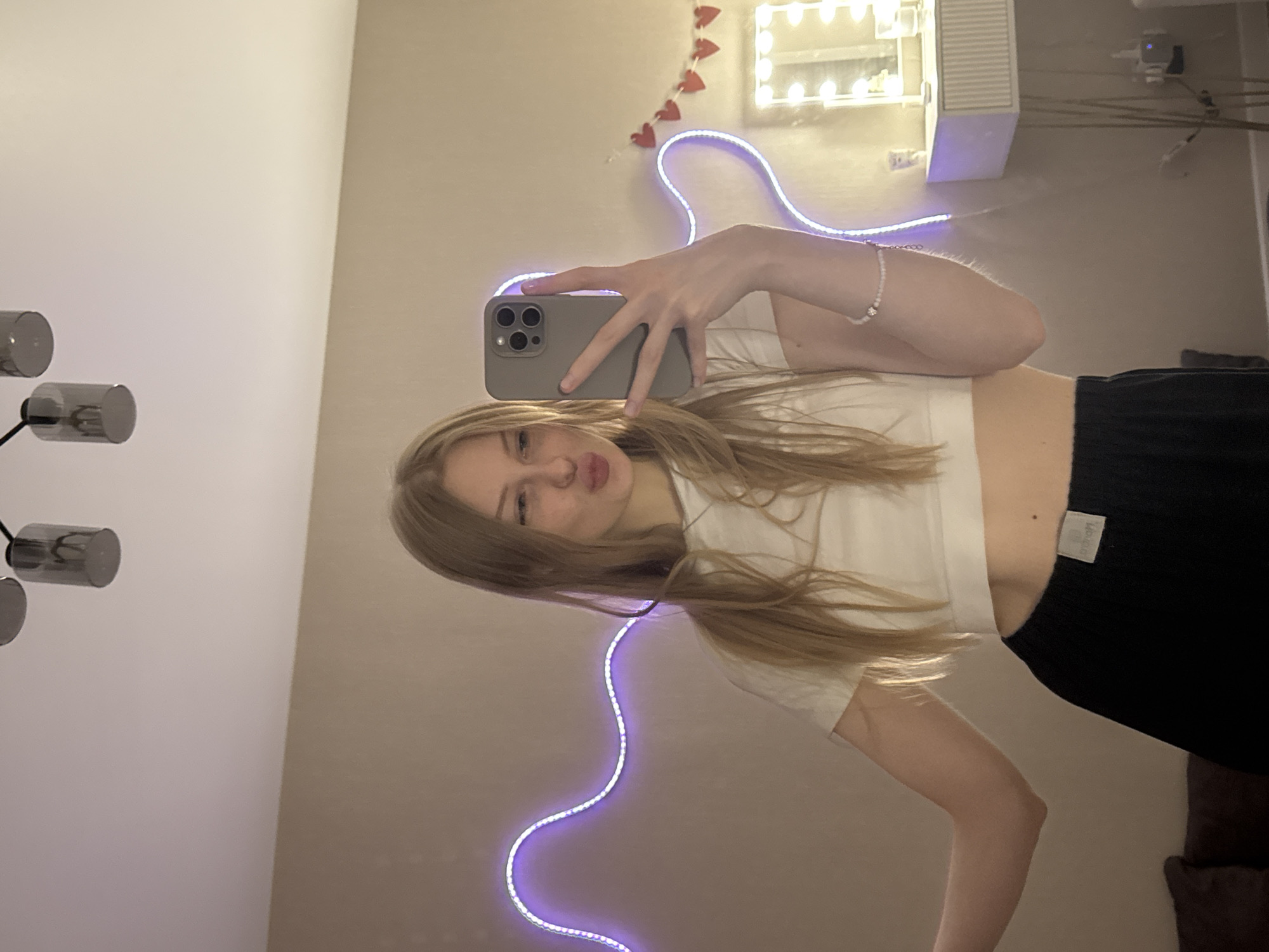 A young woman with long, straight, light brown hair is taking a mirror selfie indoors. She holds a grey-cased smartphone with her right hand, positioned in front of her face. Her expression features a slight pout and partially closed eyes, conveying a playful or casual mood. She is dressed in a white short-sleeved crop top, revealing her midriff, and dark-colored bottoms, possibly shorts or pants. A small white label with partially legible text, possibly a brand name, is visible on the waistline of her dark bottoms. A light-colored beaded bracelet is worn on her right wrist.
The background, visible in the mirror's reflection, depicts a light-colored wall adorned with various lighting and decorative elements. Above her reflected head, a rectangular vanity mirror with multiple light bulbs around its frame provides bright illumination. To the left of this vanity mirror, a string of red heart-shaped decorations is affixed to the wall. Below the vanity mirror and extending across the wall is a purple LED strip light arranged in a wavy, serpentine pattern. On what would be the left wall of the room, a modern lighting fixture with three cylindrical, dark grey/chrome-colored lamps is partially visible. Further background details include a white air conditioning unit or ventilation grill, some cables, and a dark piece of furniture (possibly a couch armrest) in the lower right corner. The artificial lighting suggests the photograph was taken indoors, likely in the evening or at night, within a contemporary room, possibly a bedroom or dressing area in Perm, Russia.