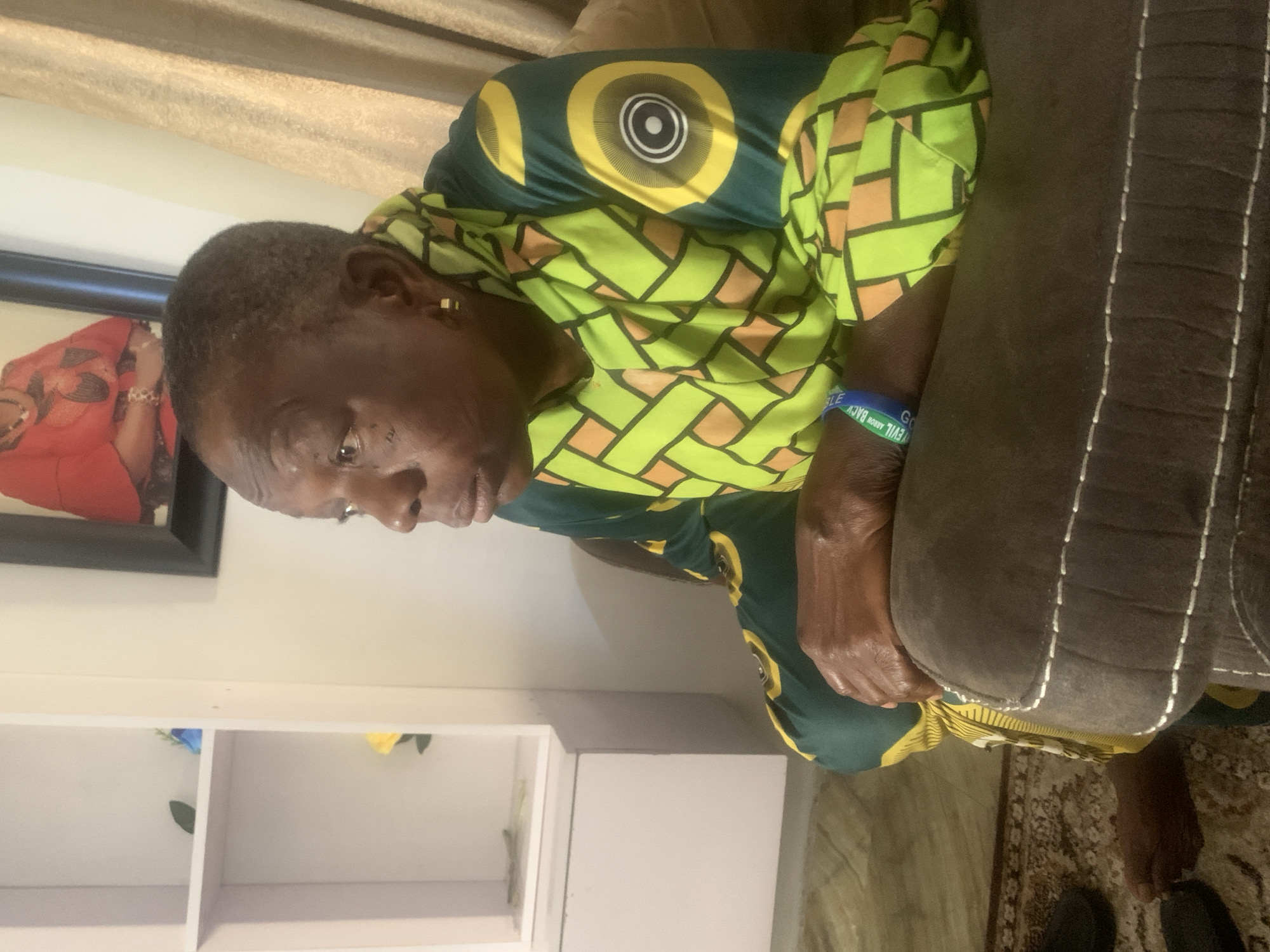 An indoor scene depicts an older adult, likely a woman, seated on a dark grey or brown sofa with prominent white stitching. The woman has dark skin and short, greying hair, and she is looking downwards and slightly to her left with a neutral, contemplative expression.
She is wearing a vibrant, short-sleeved top with a bright green base, adorned with yellow and orange geometric interlocking patterns, and large circular yellow designs on the shoulders and upper back. Small gold stud earrings are visible. On her right wrist, she wears a blue silicone wristband with white text that reads "GLE GO EVIL ARROW BACK".
The background features a light-colored wall. To the left, a framed picture shows a person, possibly a woman, dressed in a red outfit. Below the picture, a white shelving unit or cabinet is visible, with small blue and yellow artificial flowers placed on separate shelves. A patterned rug with intricate designs covers part of the floor. Above, a beige curtain suggests a window or opening. The ambient lighting indicates it is daytime. This scene is set in Jos, Nigeria.
