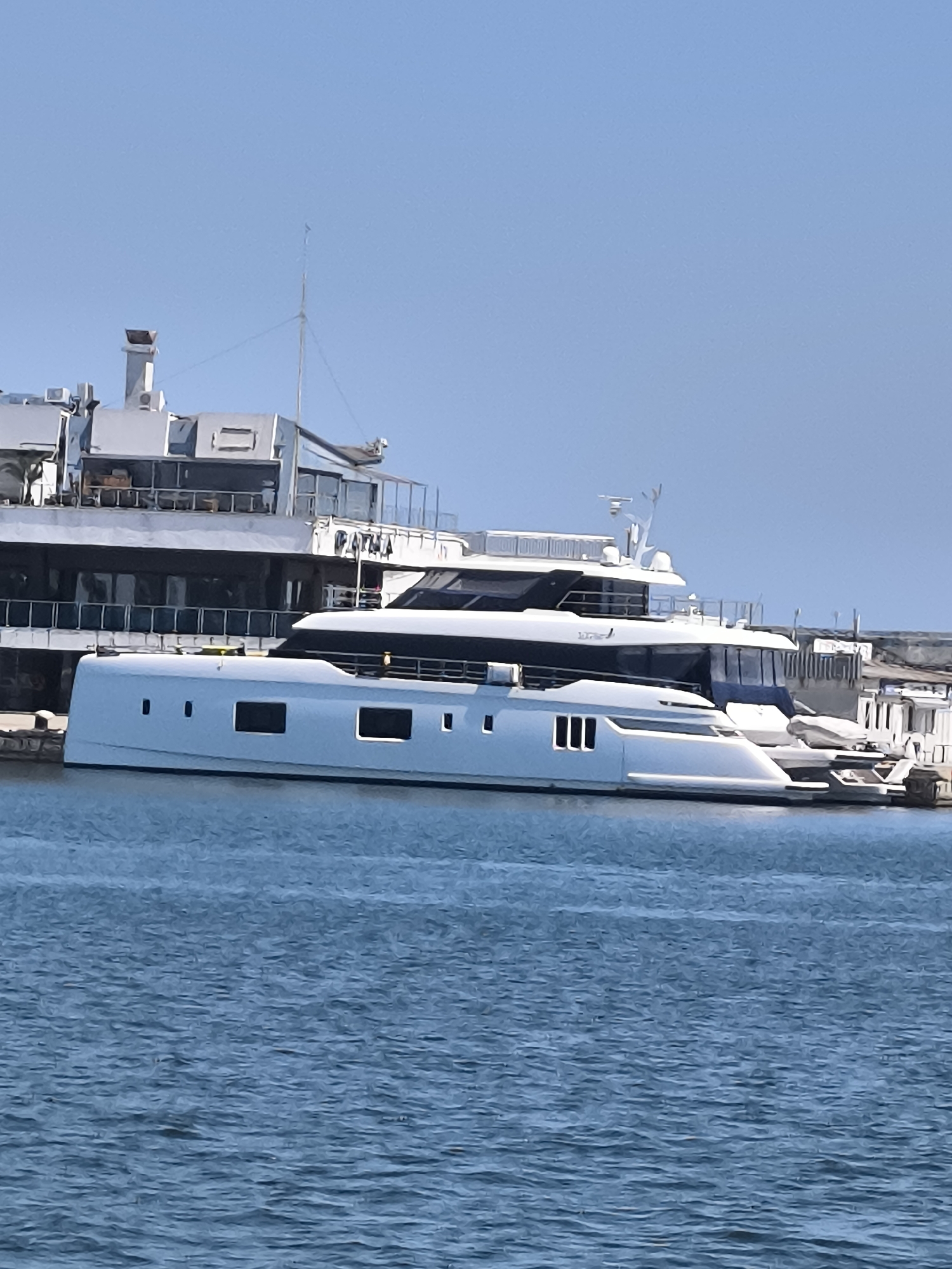 A white, modern catamaran-style yacht is docked in the foreground of a tranquil blue body of water. The yacht features a sleek, elongated hull with multiple rectangular windows. Behind it, a larger, multi-story vessel with a light-colored superstructure is visible, docked at a port facility. The background shows a clear, pale blue sky and hints of buildings and structures associated with a marina or harbor. The scene suggests a bright, sunny day. This image was captured in the Varna, Bulgaria.
