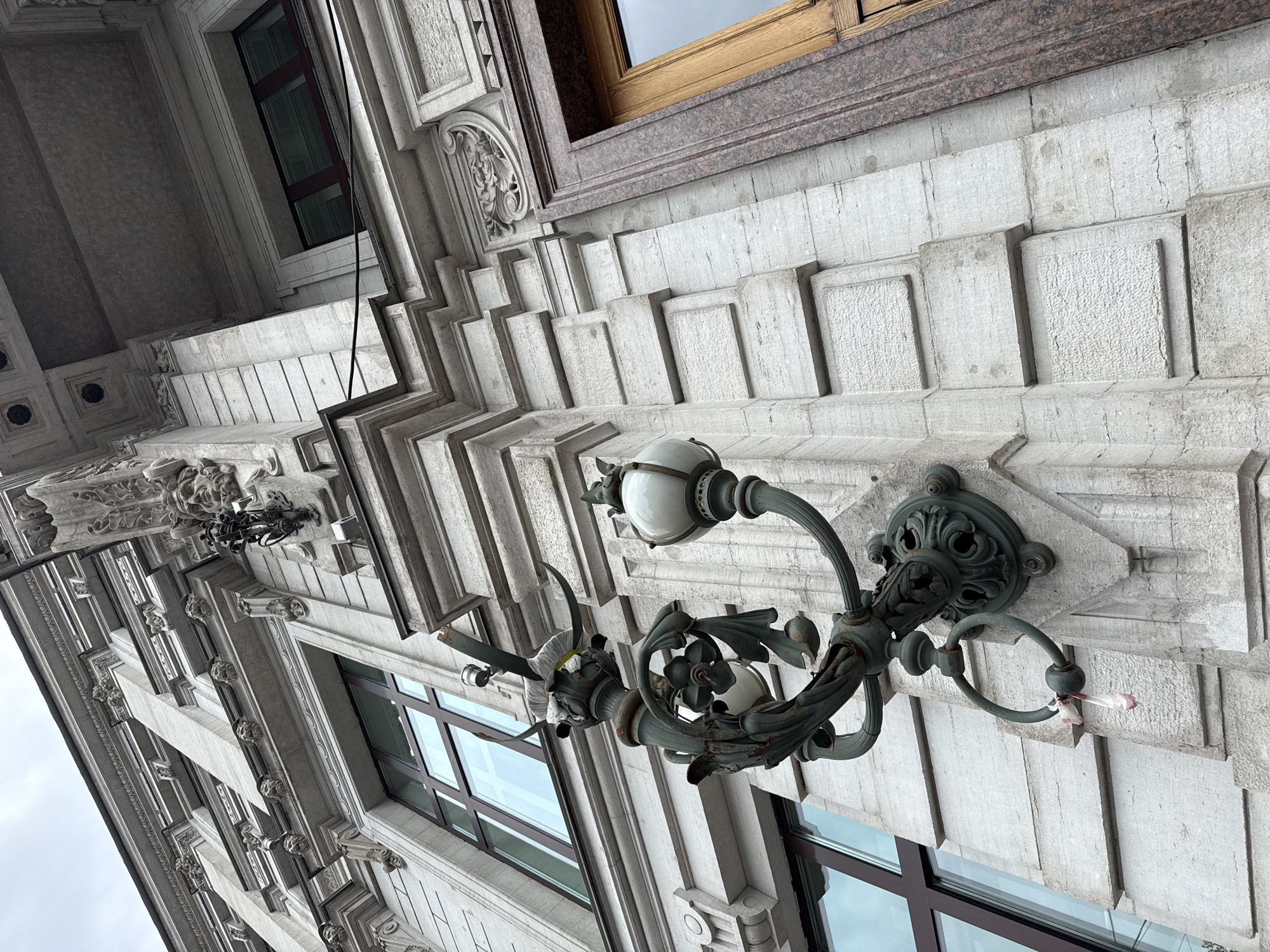 The image shows a close-up, low-angle view of the ornate facade of a building, likely in Saint Petersburg, Russia, due to the architectural style. The building's exterior is constructed of light-colored stone with detailed carvings and decorative elements. Prominently featured is a vintage-style wrought-iron lamp fixture mounted on the wall. The lamp has two globes, one of which appears to be covered with a white cloth secured by yellow tape. A small pink object, possibly a ribbon or flower petal, hangs from a ring on the lamp fixture. The building has large windows with dark frames and a section of a wooden door with a granite surround is visible on the right side of the frame. The sky above is overcast and appears grey.