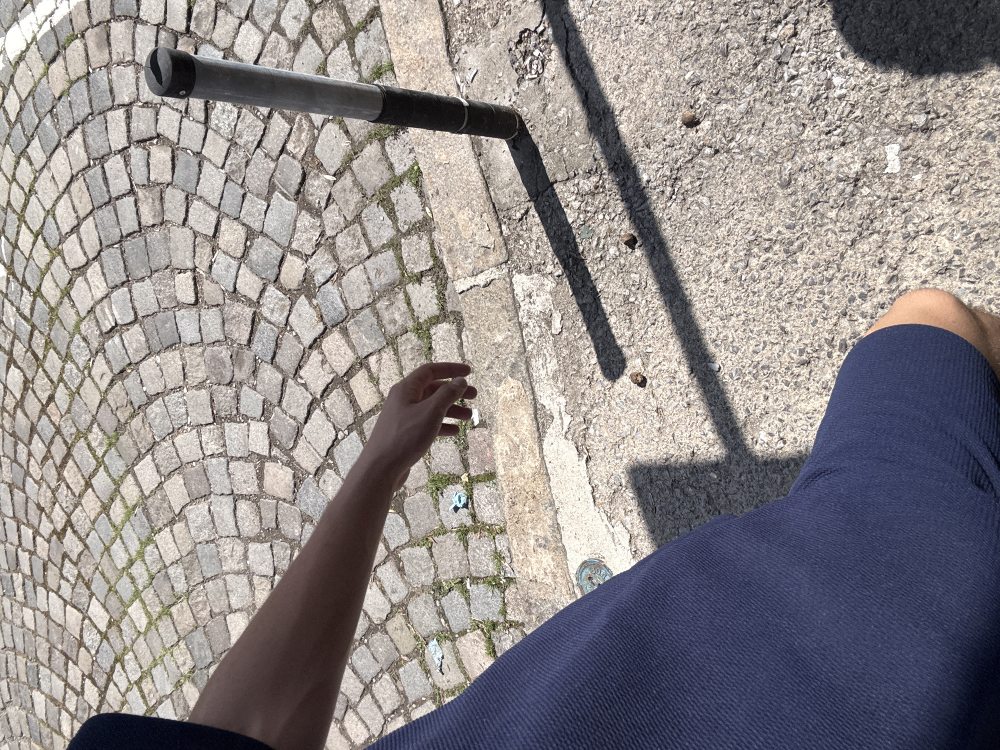 An outdoor, downward-angled photograph captures an urban groundscape under direct sunlight, featuring a partial view of an individual.
The upper-left and central portion of the image displays a pavement constructed from light-colored, irregularly shaped cobblestones, arranged in concentric, fan-like arcs. Sparse green vegetation protrudes from the interstices between these stones. A raised, rough-hewn stone curb delineates the boundary between the cobblestone pavement and an adjacent, granular, light gray concrete surface which occupies the lower-right portion of the frame. This concrete surface exhibits minor textural variations and scattered small dark particles.
A slender, metallic cylindrical bollard stands vertically on the concrete sidewalk on the right side of the image. Its upper two-thirds are light gray, while its lower third is encased in a dark, possibly black, resilient material. The bollard's apex is capped by a black, slightly convex element with a visible central groove.
Two crumpled, light-colored objects, consistent with discarded wrappers or paper, lie on the cobblestone pavement near the curb. Several small, dark, pebble-like objects are scattered across the concrete sidewalk. Partially visible in the bottom-left corner, embedded in the concrete, is a circular, dark metallic object, possibly a small utility cover or ground marker.
A bare, tanned human forearm extends from the upper-left frame margin, angled downwards and rightwards. The hand is partially visible, with fingers slightly flexed, positioned above the cobblestone surface. The lower-left portion of the image shows a segment of a human leg, clad in dark blue fabric exhibiting a fine, parallel ribbed texture. A small area of exposed skin, consistent with a knee or upper thigh, is visible at the bottom of the frame.
The scene is illuminated by direct sunlight, casting distinct, elongated shadows from the bollard and an unseen source onto the concrete sidewalk.
The radially arranged cobblestone pavement and urban infrastructure depicted are characteristic of pedestrian zones and historic city centers within Sofia, Bulgaria, commonly found in areas such as Vitosha Boulevard, Sveta Nedelya Square, or the Largo complex.