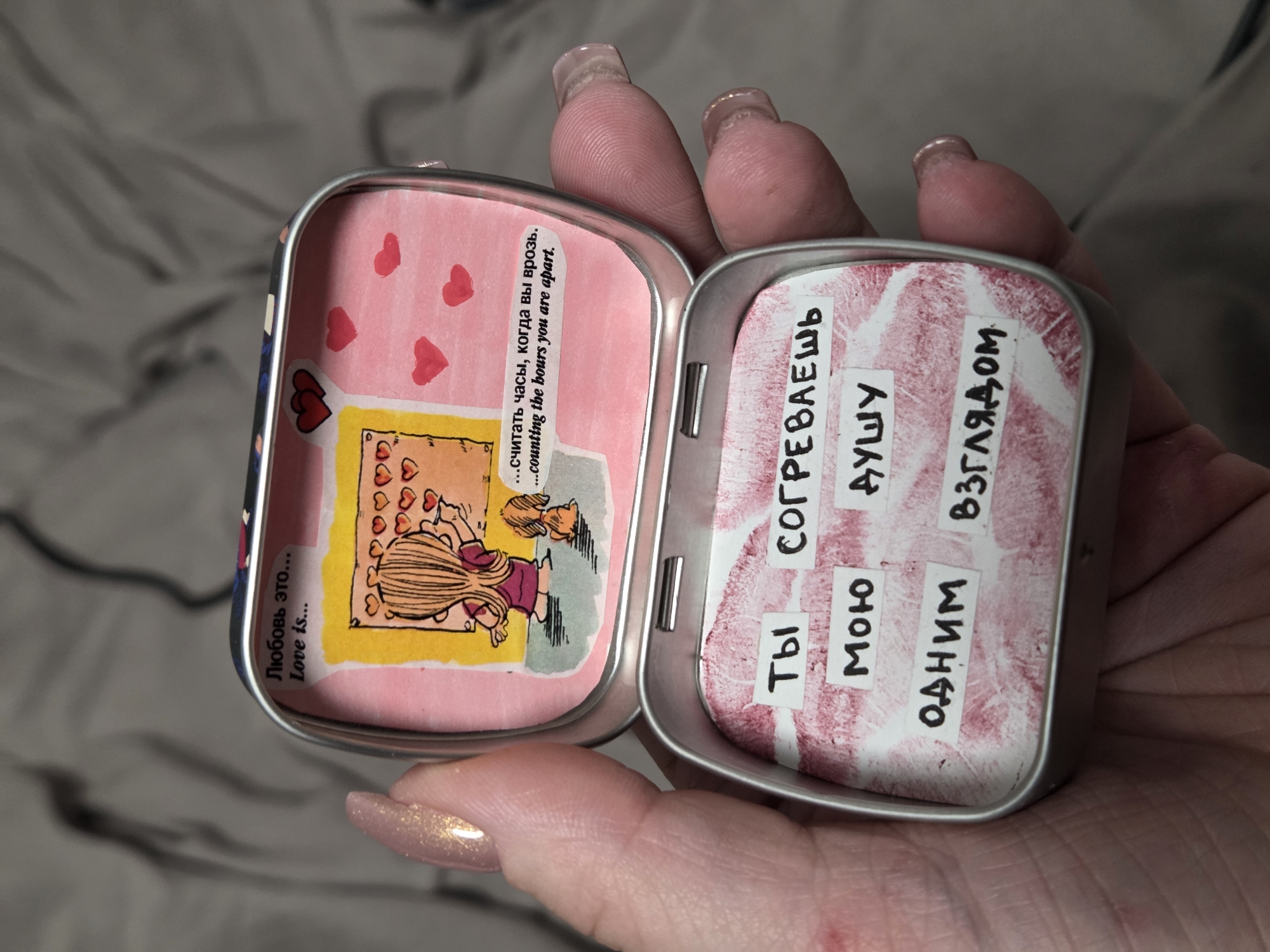 A close-up shot depicts a hand holding open a small, tin container. The container's interior is decorated with images and text. The left side shows a cartoon-style drawing of a girl and a dog, with hearts drawn above them, set against a pink background. Text in both Russian and English reads, "...считать часы, когда вы врозь. ...counting the bours you are apart." The phrase "Любовь это... Love is..." is also visible. The right side features words written on white labels against a pink backdrop resembling a lipstick stain. The text, in Russian, reads, "Ты," "СОГРЕВАЕШЬ," "Мою," "ДУШУ," and "Одним взглядом." The environment surrounding the hand and tin is out of focus and appears to be a textured, grey fabric.