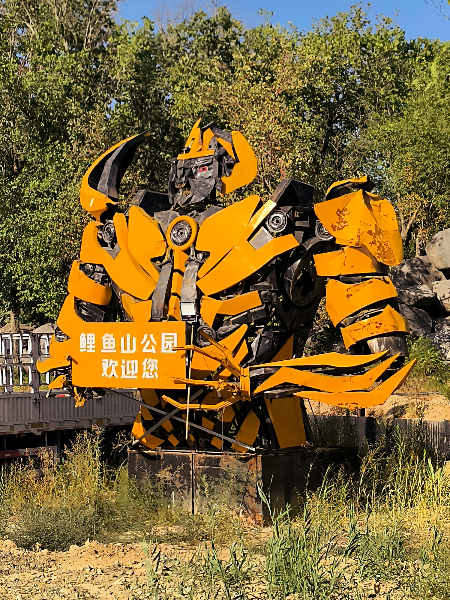 The image shows a large, yellow and black robot sculpture resembling Bumblebee from the Transformers franchise, positioned outdoors. The robot is predominantly yellow with black accents and has a menacing red-eyed face. It is holding a rectangular sign in white Chinese characters which reads "鲤鱼山公园 欢迎您" (Lǐyúshān Gōngyuán, Huānyíng Nín), translating to "Carp Mountain Park, Welcome". The sculpture appears to be made of scrap metal.
Behind the robot, there is lush green foliage, likely trees and bushes, with a clear blue sky visible above. To the left of the robot, a portion of a white truck or trailer is visible, along with a black metal fence or railing. The ground in the foreground is a mix of dry soil and green vegetation, suggesting an outdoor park or natural setting. The lighting indicates it is daytime, likely sunny, casting shadows from the robot and foliage.
While the image itself does not explicitly state the location in Ürümqi, China, the presence of Chinese text on the sign strongly suggests it is within China. The name "Carp Mountain Park" (Lǐyúshān Gōngyuán) is a geographical indicator. Research indicates that Lǐyúshān Gōngyuán is a park located in the Tianshan District of Ürümqi, Xinjiang, China. Therefore, this sculpture is likely located within Lǐyúshān Gōngyuán in Ürümqi, China. The scene depicts a static display, likely a tourist attraction or decorative element within the park. No people are visible in the image.