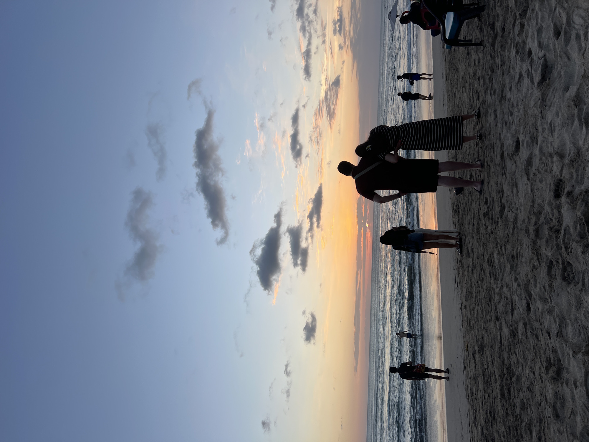 An aerial view of Kuta Beach in Kuta, Indonesia, shows a group of people enjoying the sunset. The sky is filled with soft hues of orange and pink, with scattered clouds. The ocean waves gently lap onto the sandy shore, where silhouettes of people can be seen. In the foreground, a couple stands embracing, facing the ocean. Further down the beach, individuals are walking and wading in the water. The scene evokes a sense of tranquility and natural beauty.