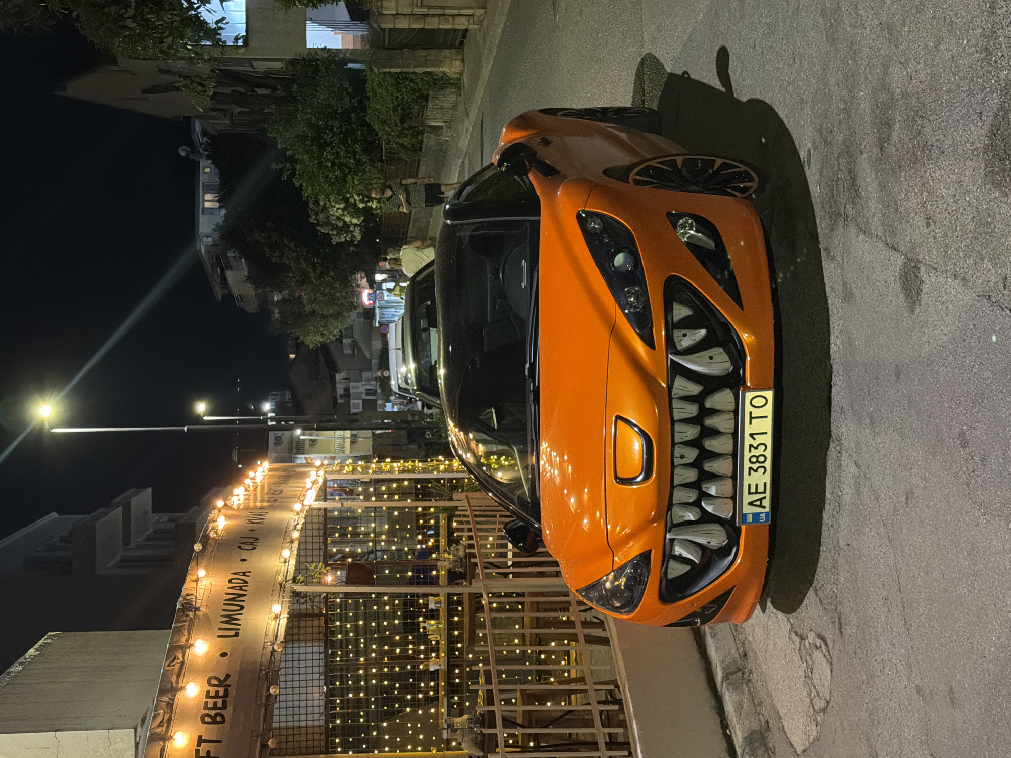 An orange sports car with a monstrous grille, resembling sharp teeth, is parked on an asphalt street at night. The car's headlights are on, illuminating the street. In the background, a building with a sign that reads "CRAFT BEER" and "LIMUNADA" is decorated with numerous string lights, suggesting it is a restaurant or bar. Two individuals are visible in the distance, walking away from the car. One is wearing a dark patterned shirt and shorts, and the other is wearing a light-colored t-shirt and pants. The street is lined with buildings and trees, and a streetlight casts a beam of light downwards. The license plate of the car reads "AE 3831 TO" and has a Ukrainian flag emblem. The scene appears to be in the city of Budva, Montenegro, as indicated by the presence of a sign for "LIMUNADA" (lemonade), which is a common offering in the region.