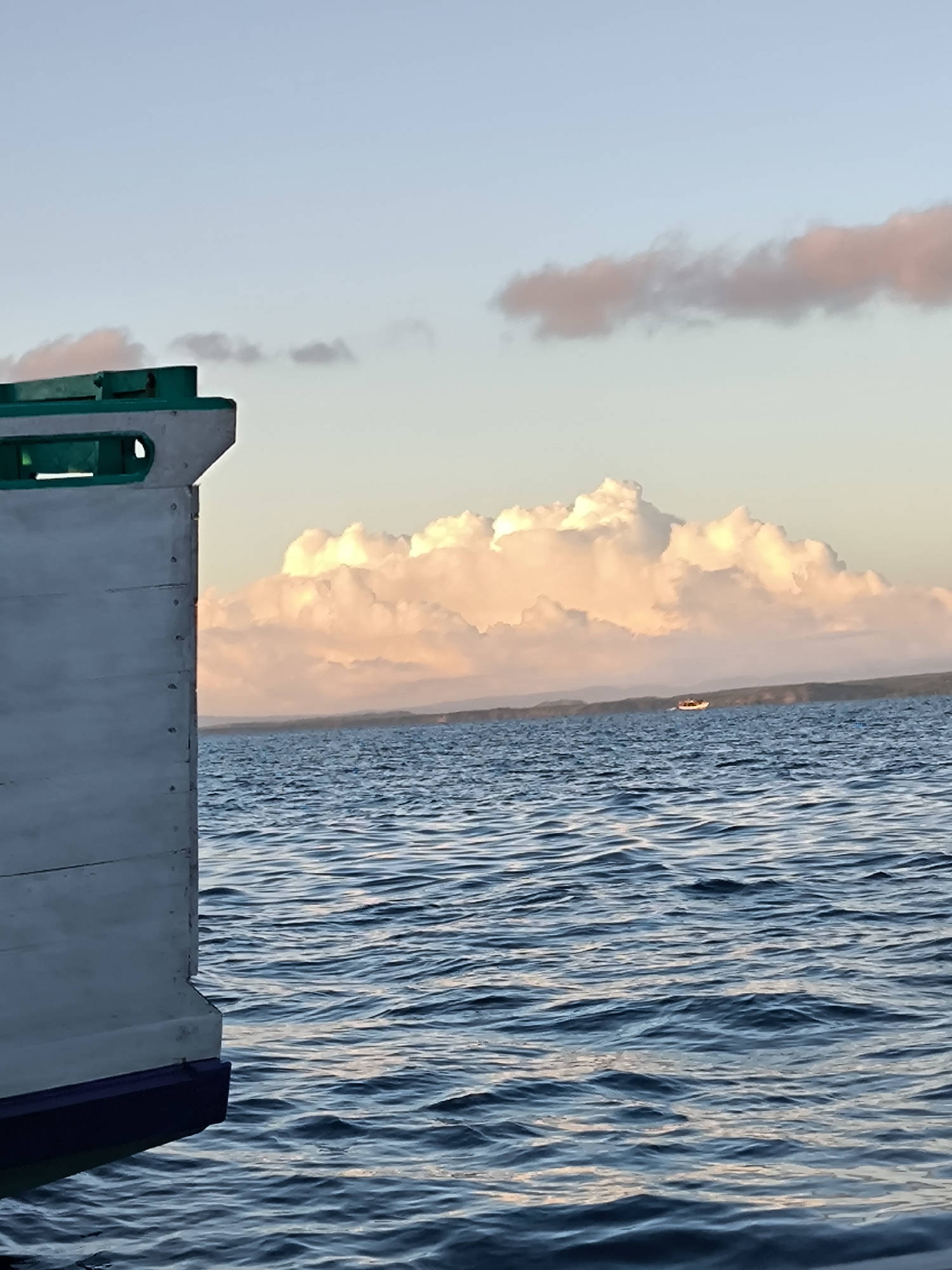 The image depicts a marine scene in Unknown, Indonesia. The foreground shows a portion of a boat, painted white with teal and purple accents. The midground features a body of water with gentle waves, and a small boat is visible in the distance. The background consists of land, a large cumulus cloud lit by the setting sun, and a pale blue sky with faint pink clouds.