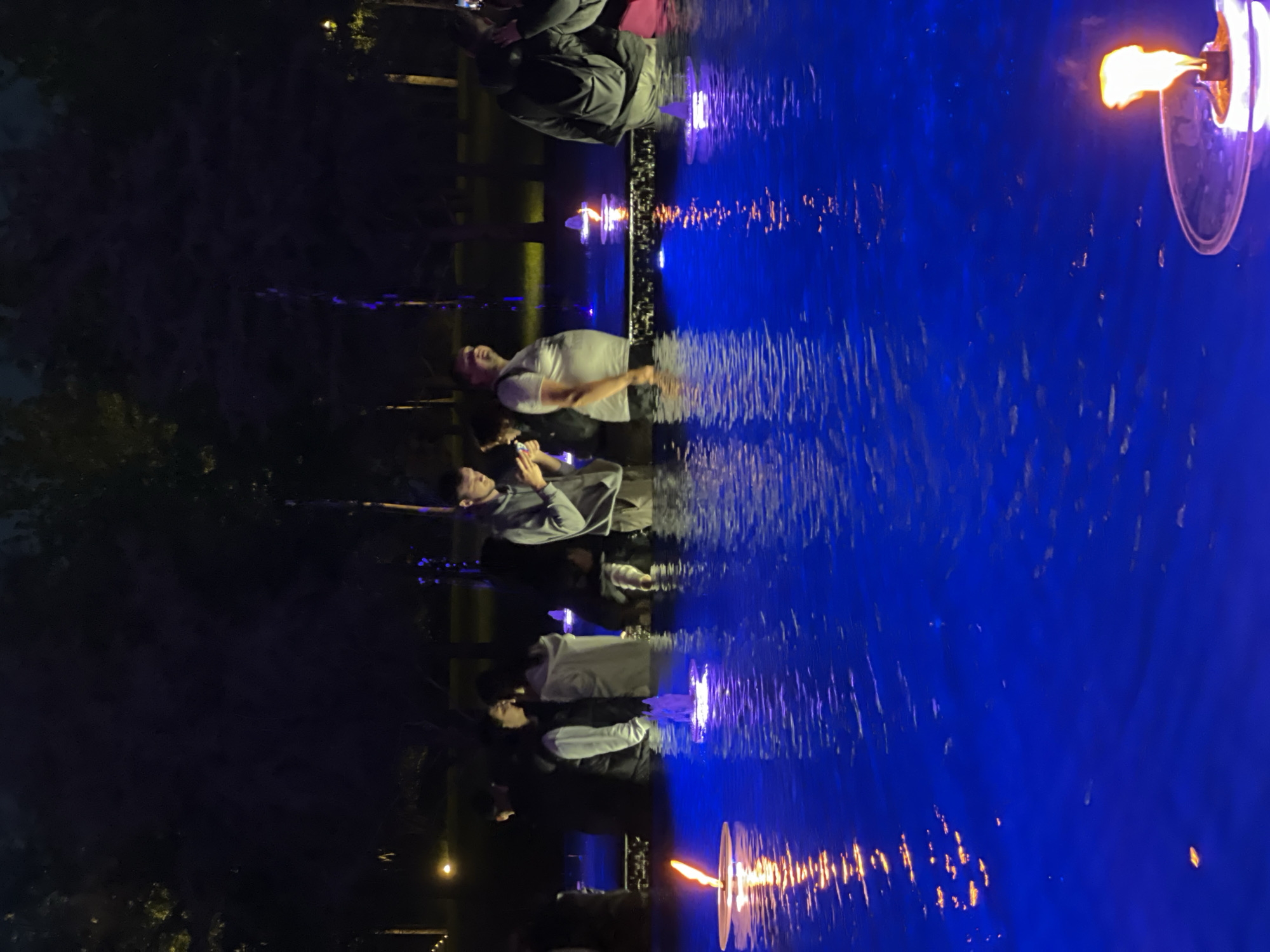 This image captures a night scene in Krasnodar, Russia, centered around a body of water illuminated by vibrant blue light. Several people are gathered on the left side, observing the water and the floating flames.
In the foreground, a man in a white t-shirt and glasses stands, looking towards the water. Below him, another person in a grey jacket is seated on an edge, engaged with a smartphone. Further to the left, more individuals are visible, some seated along what appears to be a low wall or embankment. The background on the far left is dark, hinting at trees or foliage against the night sky.
The most striking feature is the water, which occupies the right half of the frame and glows with an intense blue hue. Its surface is gently rippled, reflecting the blue light and the warm, orange-yellow glow of several floating fire elements. These elements resemble floating candles or torches, with visible flames dancing above the water. One large floating flame is prominent in the upper right, another in the lower right, and a smaller one is seen closer to the observers.
The scene conveys a calm and contemplative atmosphere, suggesting a public event, art installation, or a decorative display in a park or waterfront area during the evening or night. The primary light sources are the blue illumination of the water and the natural fire from the floating elements. No text is visible in the image.