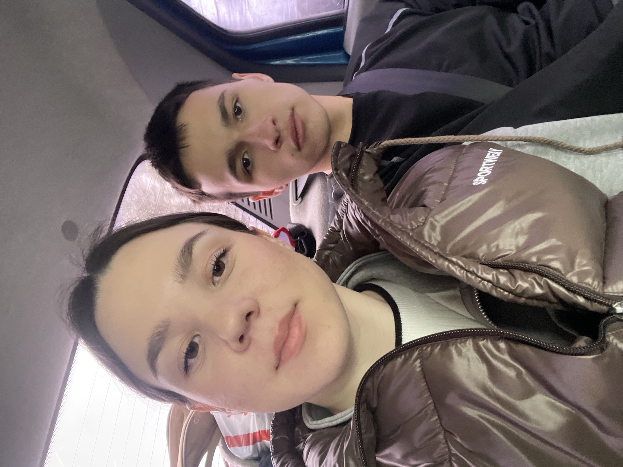 Two young adults are in a vehicle. The woman is in the foreground wearing a brown, shiny puffer jacket over a gray hoodie and a white shirt with a black stripe. The man is behind her, wearing a black shirt and a seatbelt. The interior of the vehicle is visible, including the window and a blue panel. It is not possible to determine a specific location within the city of Uspenskii, Russia from the provided image.