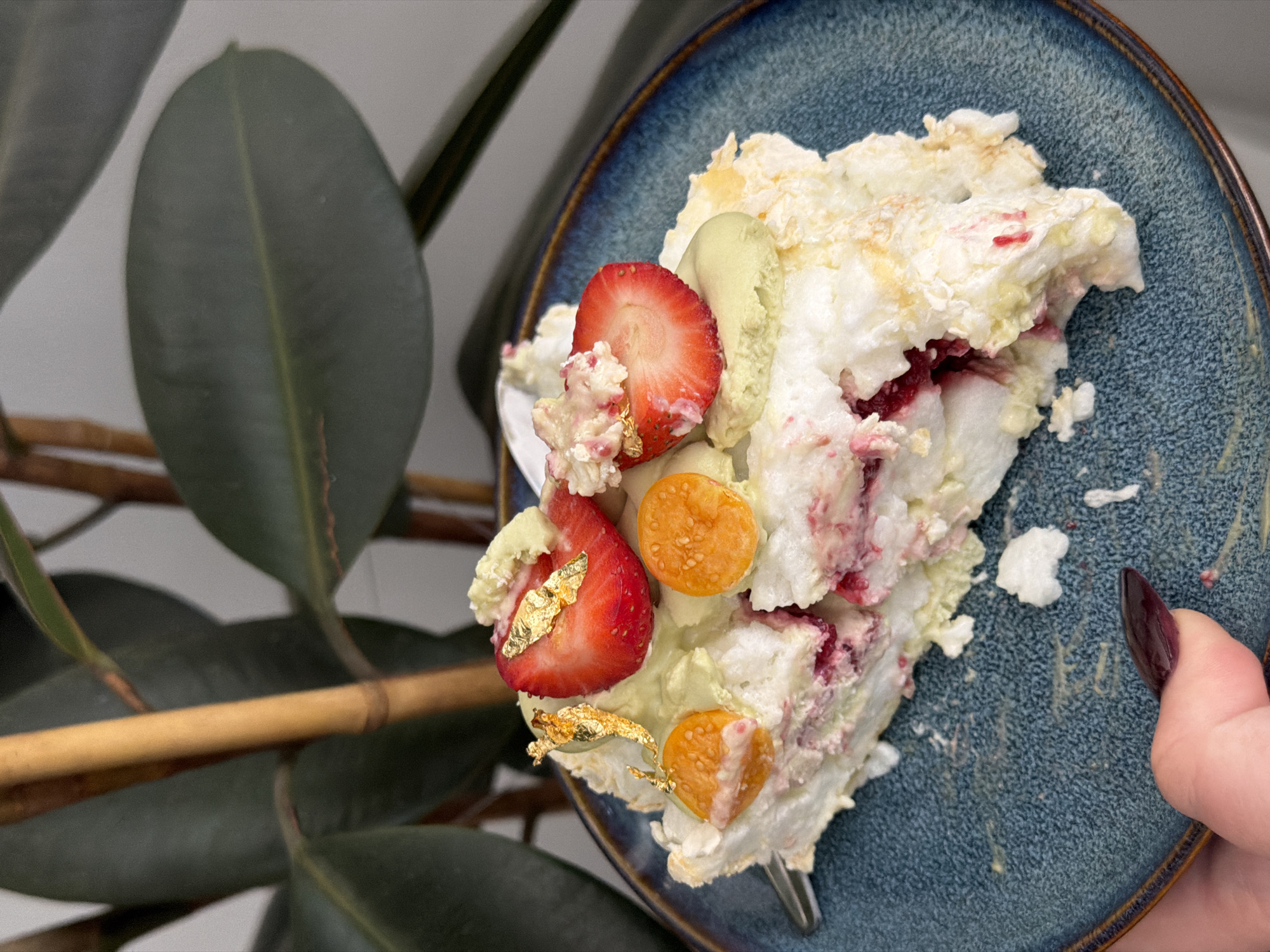 A plate of food, consisting of white meringue-like material, strawberries, golden berries, and gold leaf, sits on a blue ceramic plate with a brown rim. A hand with a long, dark fingernail is holding the plate. In the background, there's a plant with large, dark green leaves and a light brown stem. The location is Ramat Gan, Israel.