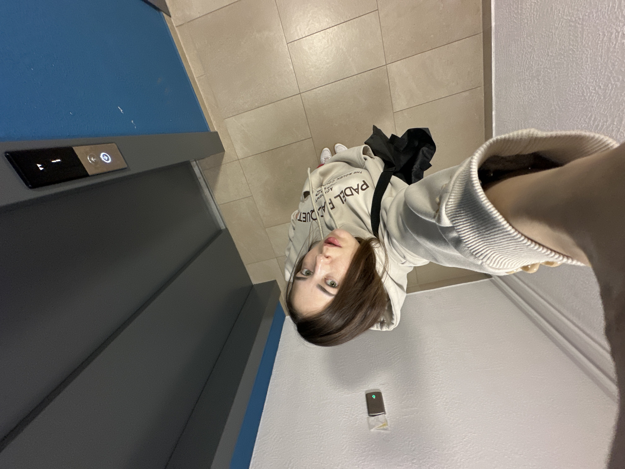 The image shows a young woman in a light-colored hoodie and white sneakers, holding a black bag over her shoulder. She is standing in what appears to be a residential building hallway, with beige tiled flooring and light grey and blue walls. To her left, there is a grey elevator door with a panel of buttons, including illuminated up and down arrows and a circular button with an illuminated door icon. To her right, on a textured white wall, is a small, dark rectangular object, possibly an access card reader or intercom, with a green illuminated light.
The woman is looking directly at the camera, her face angled slightly upwards, with a neutral expression. Her eyes are green and her hair is brown and shoulder-length. The lighting suggests it is daytime, though indoor lighting is also present. The overall impression is that she is about to enter or exit an elevator. The clothing and environment do not provide specific clues about the time of day or weather outside. The visible text on the elevator panel includes directional arrows and a door symbol. Text on her hoodie is partially obscured but includes "PADEL ACADEMY" and "THE ROUEN COU..." indicating a connection to a sports academy.