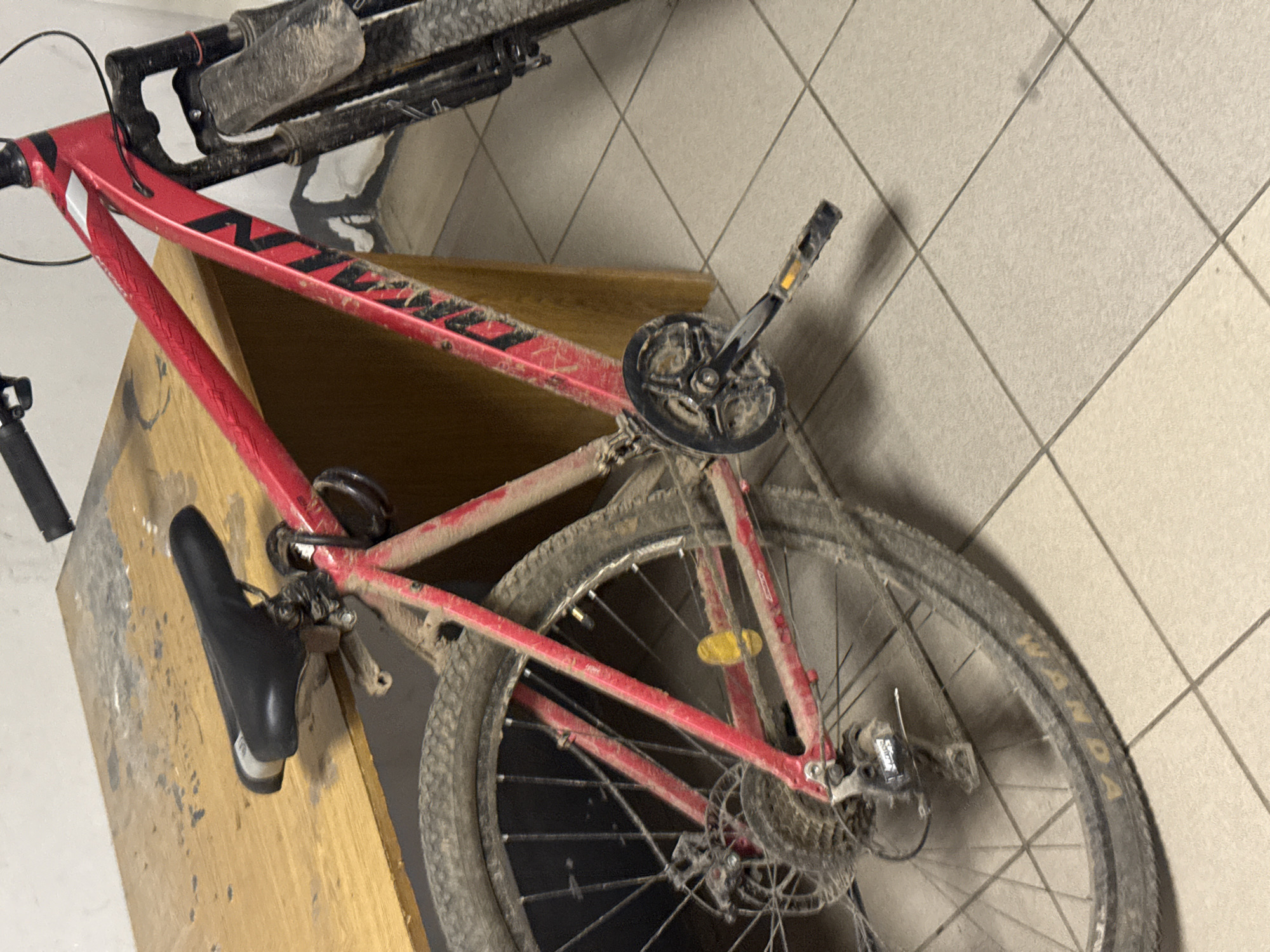 The image shows a muddy red mountain bike leaning against a wooden surface. The bike is heavily soiled, with mud covering the frame, tires, and components. The rear tire brand is partially visible, displaying "WANDA". The bike is indoors, positioned on a tiled floor. No people are visible. The location is stated to be Votkinsk, Russia but no other details confirming the location are apparent in the photograph.