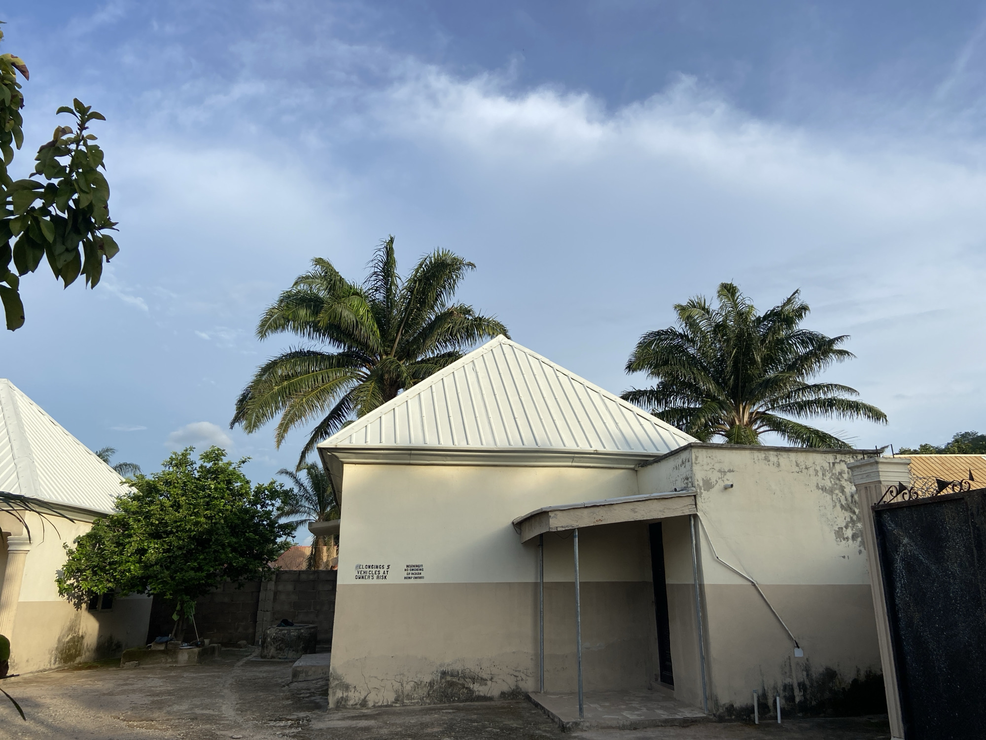 This image depicts an outdoor scene in Mararaba, Nigeria, featuring two single-story buildings with white corrugated metal roofs and two-toned walls, cream on top and beige below. The main building in the foreground has a small, covered entrance on its right. Visible on its wall are two signs: "BELONGINGS & VEHICLES AT OWNER'S RISK" and "WARNING! NO SMOKING OF INDIAN HEMP (WIWI)."
A lush green tree stands between the buildings, with another building partially visible to its left. Several tall palm trees with green fronds are prominent in the background and on the right, towering above the structures. The ground is a mix of concrete and dirt, and a black metal gate with decorative elements can be seen on the far right. The sky is partly cloudy with patches of blue, and the lighting suggests either late afternoon or early morning, casting soft shadows. No people or specific activities are present, portraying a quiet, residential or compound setting.