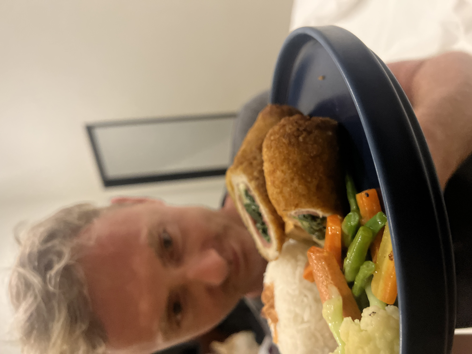 The image presents a close-up, tilted view of a meal served on a dark blue plate, with a blurred person visible in the background. The food in the foreground is in sharp focus and consists of two golden-brown, breaded rolls, one of which is cut open to reveal a filling of green leafy vegetables (likely spinach) and what appears to be ham. Accompanying the rolls is a portion of fluffy white rice and a vibrant mix of steamed or boiled vegetables, including bright orange carrots, green beans, and white cauliflower.
In the out-of-focus background, a person with wavy, light-colored hair and fair skin is partially visible, appearing to look towards the food. Their expression is indistinct due to the blur and angle, but suggests mild interest or observation. The setting appears to be indoors, possibly a room in Kuta Selatan, Indonesia, under ambient or artificial lighting. No specific time of day or weather conditions can be determined from the indoor environment. There is no visible text in the image. The overall composition suggests a personal photograph of a meal, possibly moments before being eaten.