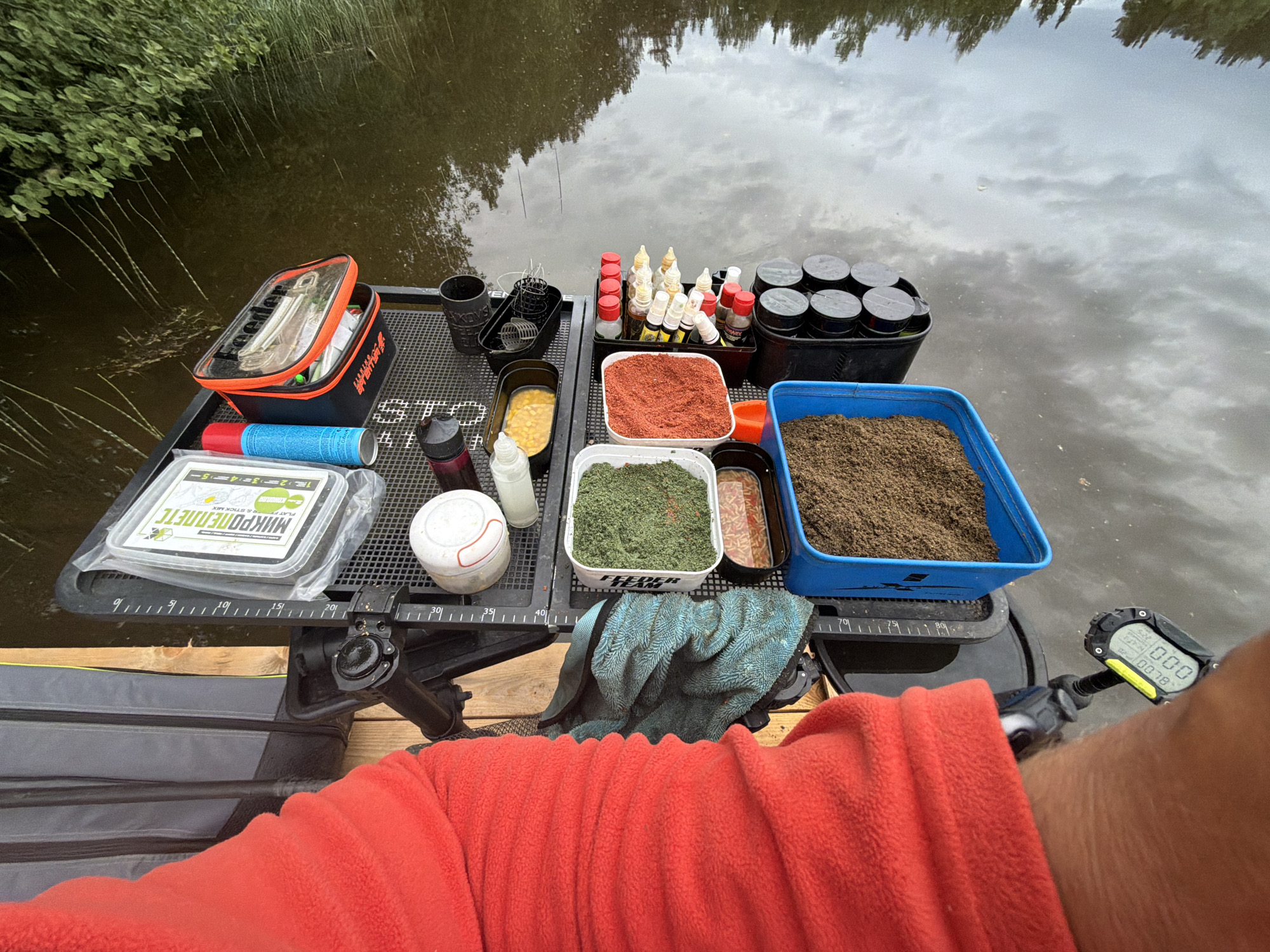 A person in a red fleece jacket is seated on a wooden platform near a body of water. On a black, mesh table in front of them is an assortment of fishing equipment and bait. The fishing gear includes a black and orange tackle box, bait containers filled with various types of bait (corn, red powder, green powder, brown soil), liquid additives in bottles, and small canisters. There is also a white container labeled "MIKROPELLETS". A blue towel is draped over the center of the table. The platform appears to be a fishing platform setup alongside a pond or river. The water reflects the sky and vegetation. The person's arm and a digital fishing device are visible on the right side of the image. The location is likely Prikamskii, Russia, based on geographical cues.