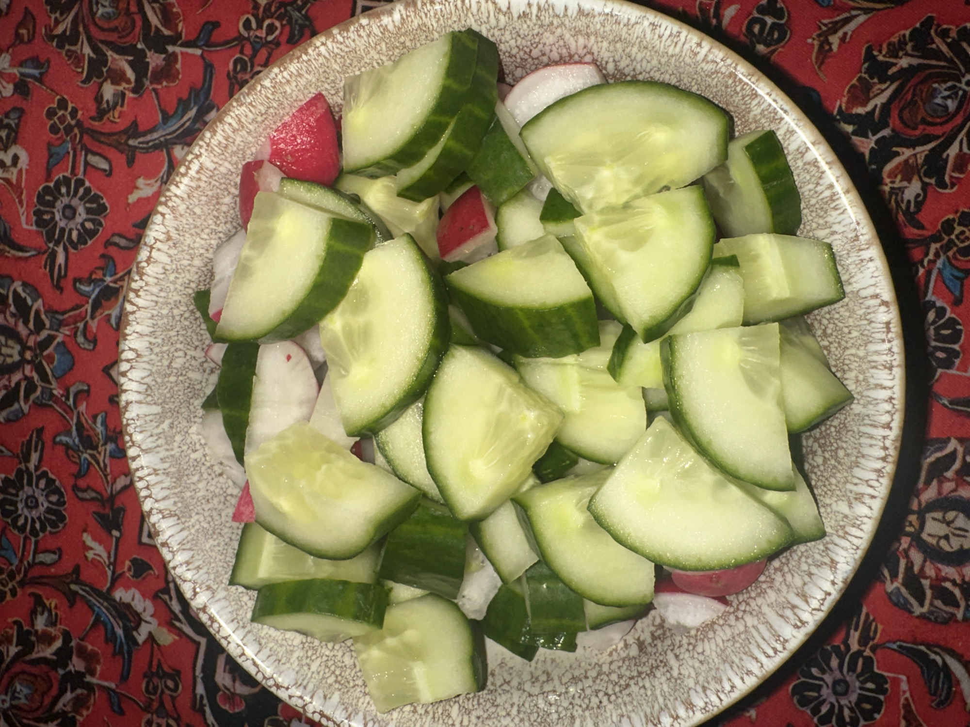 A bowl of sliced cucumbers and radishes is centered on a red patterned tablecloth. The cucumbers are pale green with darker green skins, and are cut into semi-circular slices. Red radishes are interspersed within the cucumber slices. The bowl has a mottled brown and tan rim. The tablecloth has a red background with a pattern of stylized flowers and leaves in dark red, brown, and black. The scene is in a location in the city of Blagoveshchensk, Russia.
