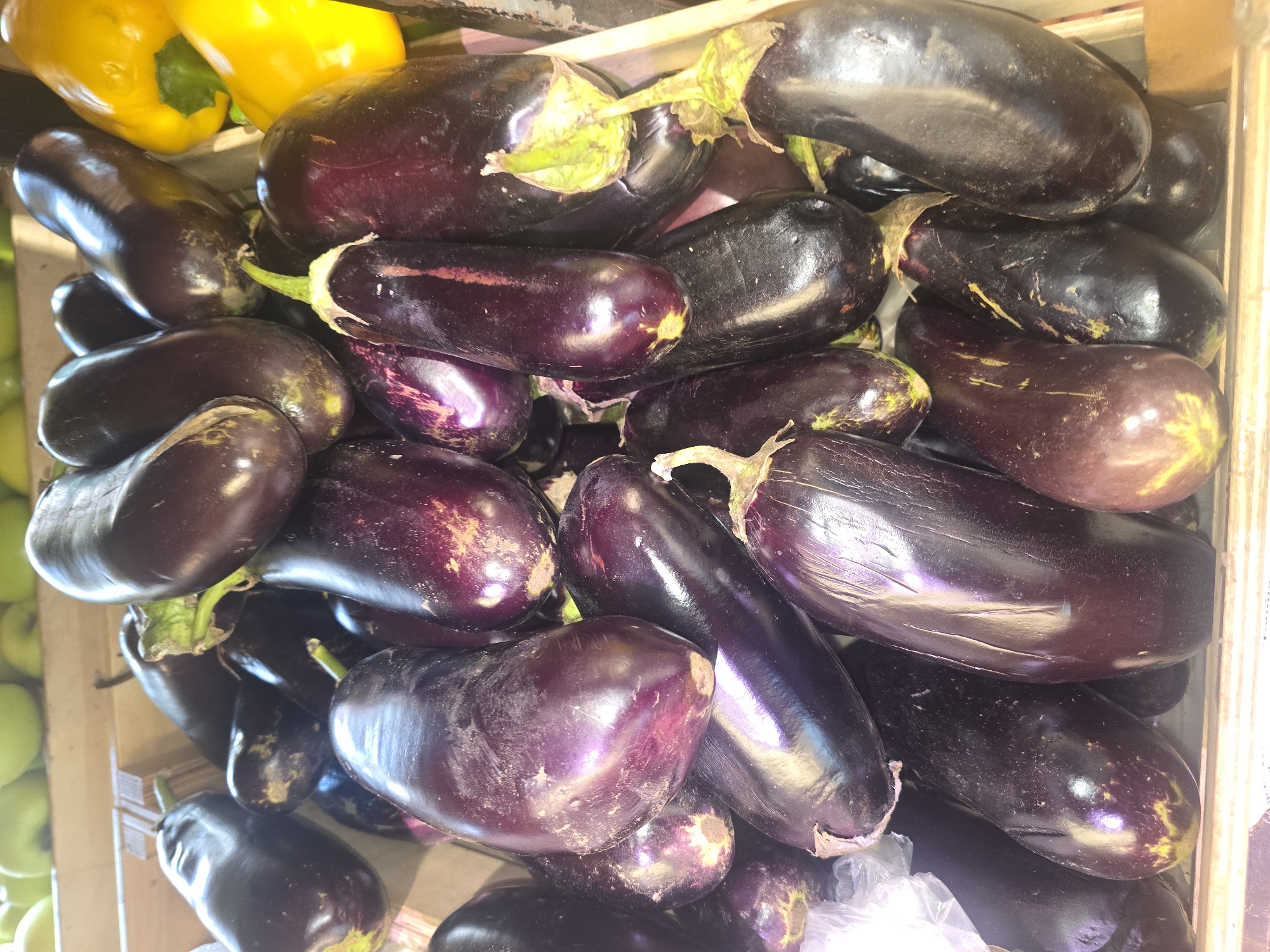 This is a close-up, overhead shot of a display of eggplants at an outdoor market in Ulan-Ude, Russia. The eggplants are primarily a deep purple color, with some having slight green and yellow hues near their stems. They are of varying sizes and shapes, with some being elongated and others more bulbous. Some of the eggplants appear to have white speckling or blemishes on their skin. The eggplants are piled into a wooden crate, with the stems and tops of some visible. In the background, a few yellow bell peppers are also visible in the crate, along with the tops of green apples. The lighting is bright, suggesting it is daytime.