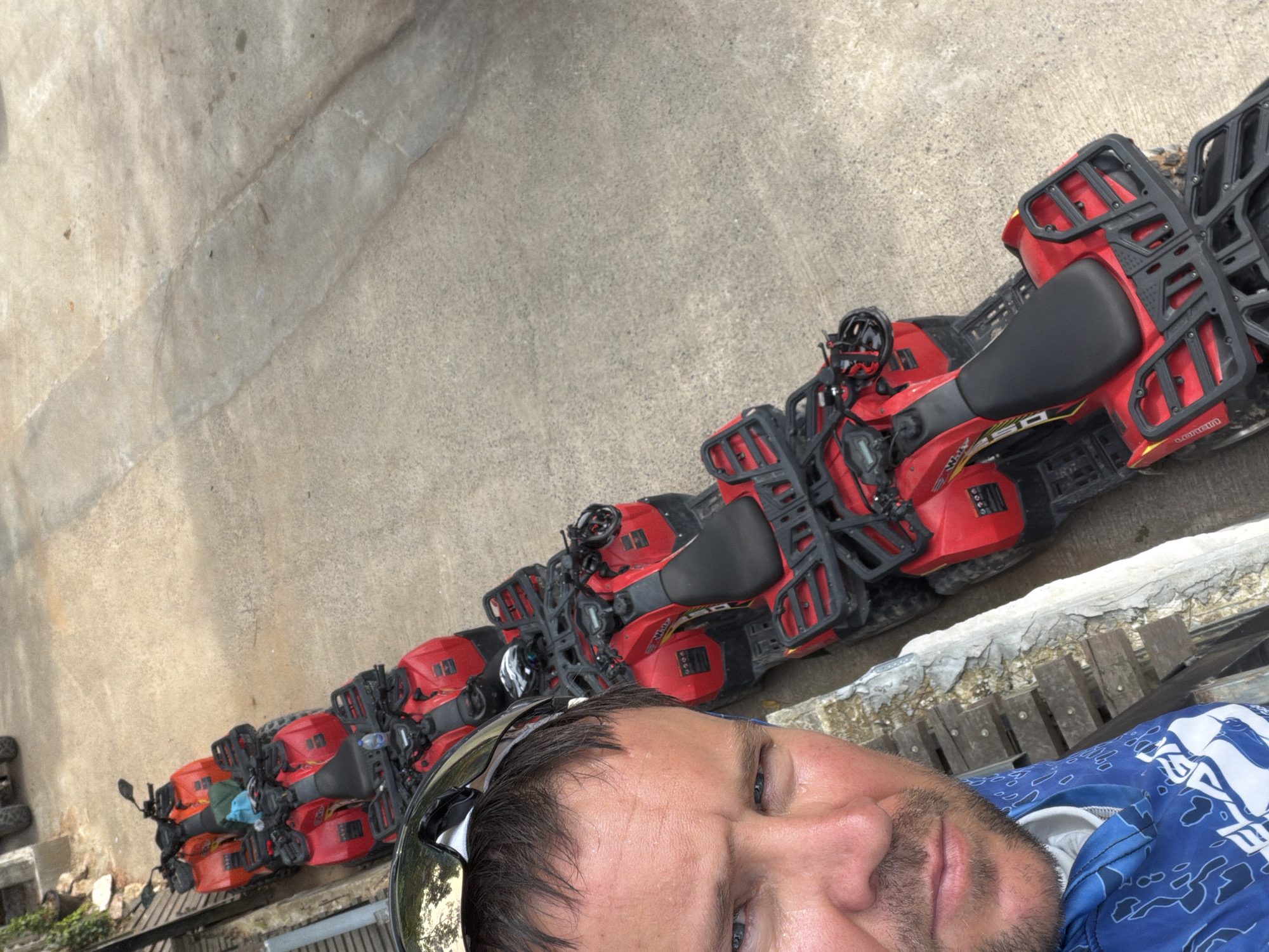 A man, with short brown hair, stubble, and blue eyes, takes a selfie while wearing sunglasses pushed up on his head and a blue patterned shirt. He is standing next to a line of red quad bikes parked on a concrete surface, with a wall and wooden planks to his left. The quad bikes appear to be part of a rental or tour operation. The scene is likely in Ko Samui, Thailand, given the context of the quad bikes and tropical foliage visible in the background. The lighting suggests it is daytime, possibly overcast.