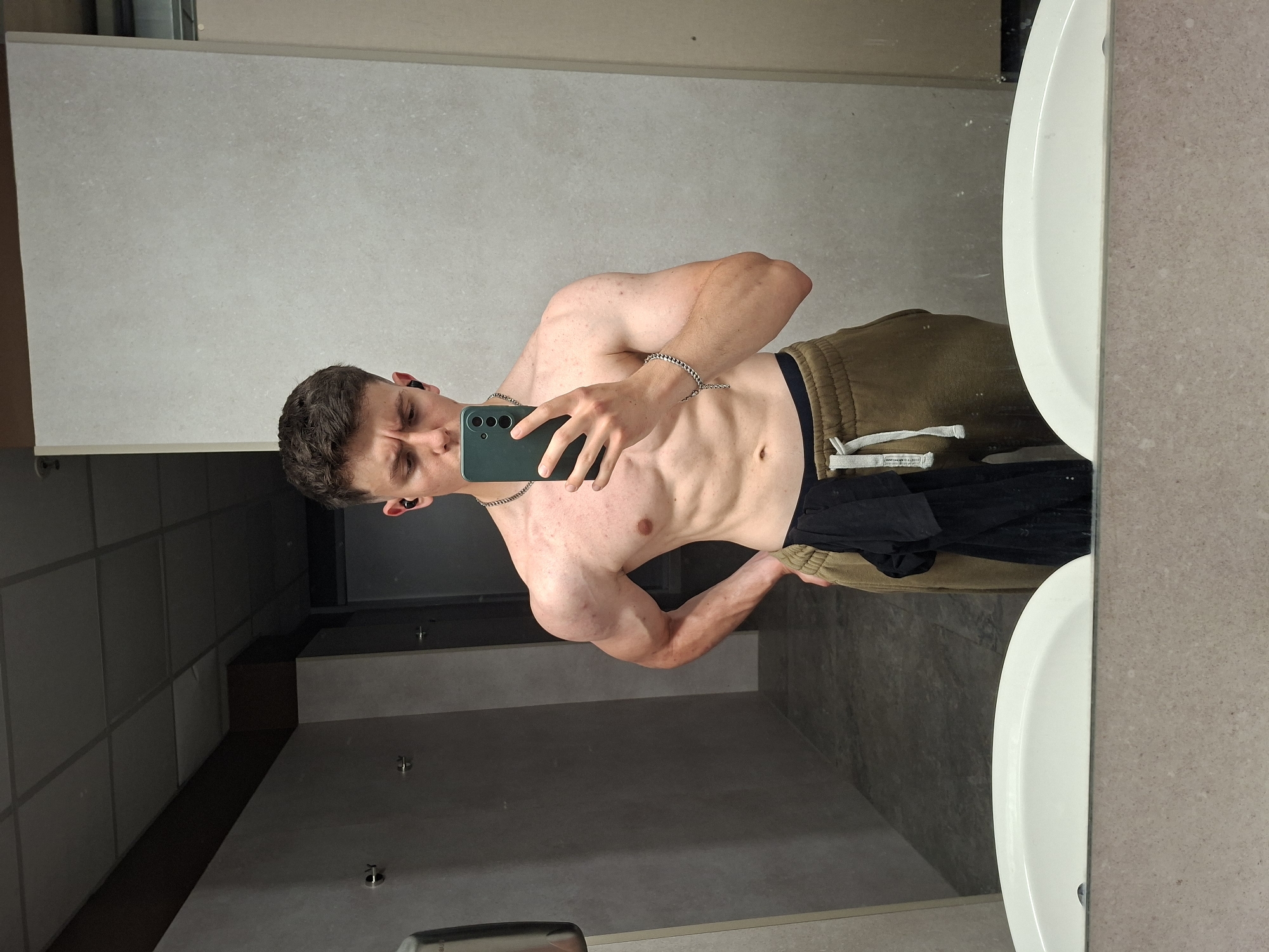 A young man takes a shirtless selfie in a bathroom mirror. He is wearing brown sweatpants with a black waistband and a silver chain necklace and bracelet. He is holding a smartphone with a green case. Black clothing is draped over his sweatpants. The bathroom has a grey tiled wall and floor, with a white sink visible in the mirror's reflection. The location is likely a public restroom, but not identifiable within the city of Plovdiv, Bulgaria.