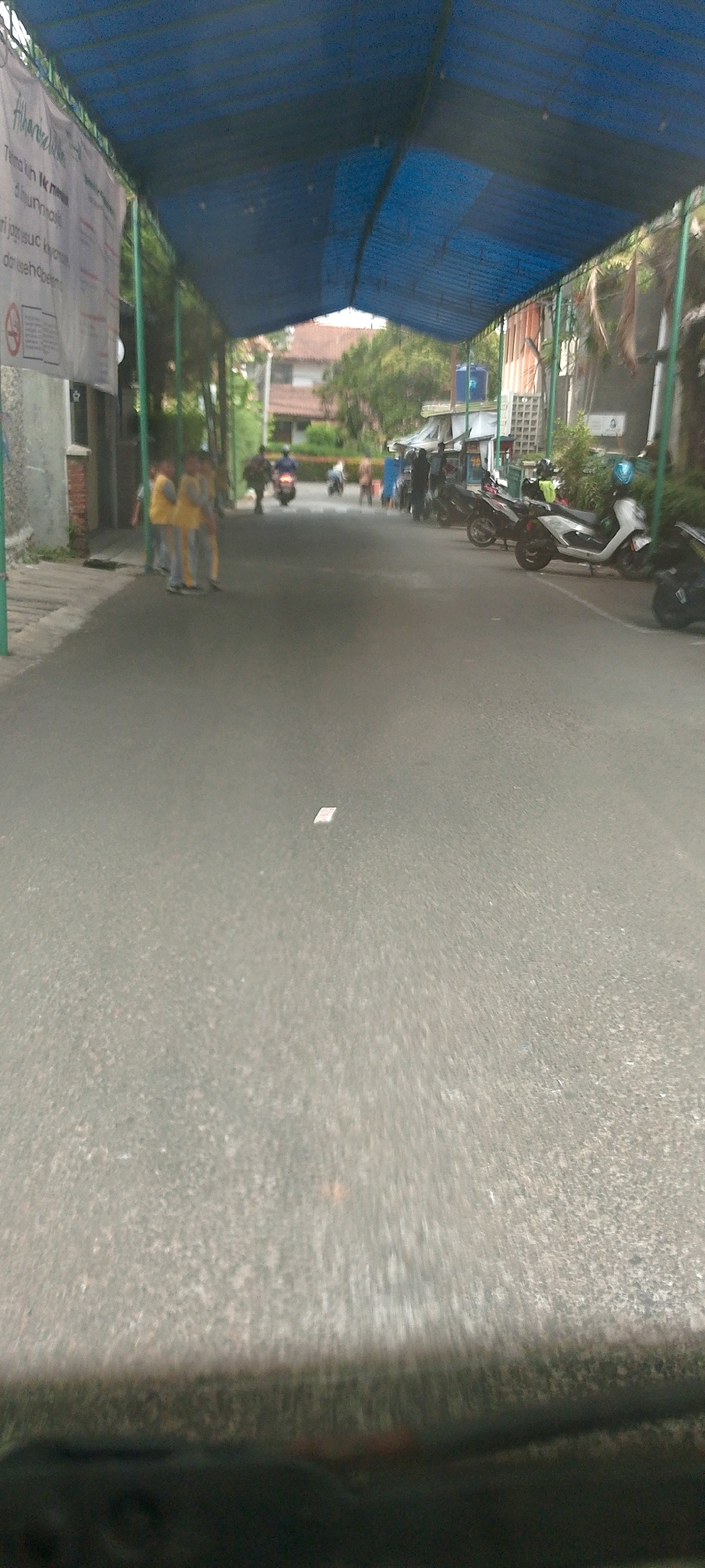 This media file captures a street scene under a blue canopy, likely in Bandung City, Indonesia. The perspective suggests the image was taken from a vehicle, as the foreground is blurred and dark, resembling a dashboard or car interior.
In the mid-ground, several people are visible. On the left side, three children in yellow and gray uniforms stand on the sidewalk. Further down the street, a group of people are gathered near some small stalls or shops, and several motorbikes are parked or in motion. One white motorbike is particularly prominent on the right side. A few other motorbikes can be seen further down the road, suggesting vehicular traffic.
The setting appears to be a somewhat commercial or residential street with buildings lining the sides, partially obscured by greenery. The blue canopy overhead casts shadows, and the lighting suggests it might be daytime, possibly overcast or late afternoon. There is a small white object, possibly a piece of paper, lying in the middle of the asphalt. A banner with text is visible on the left side of the image, though the text is largely illegible due to distance and resolution. The visible portion of the banner shows some Indonesian writing, with "humak" being the only decipherable word. The overall impression is a busy, everyday street scene in an urban environment.
--- OCR Start ---
humak
--- OCR End ---