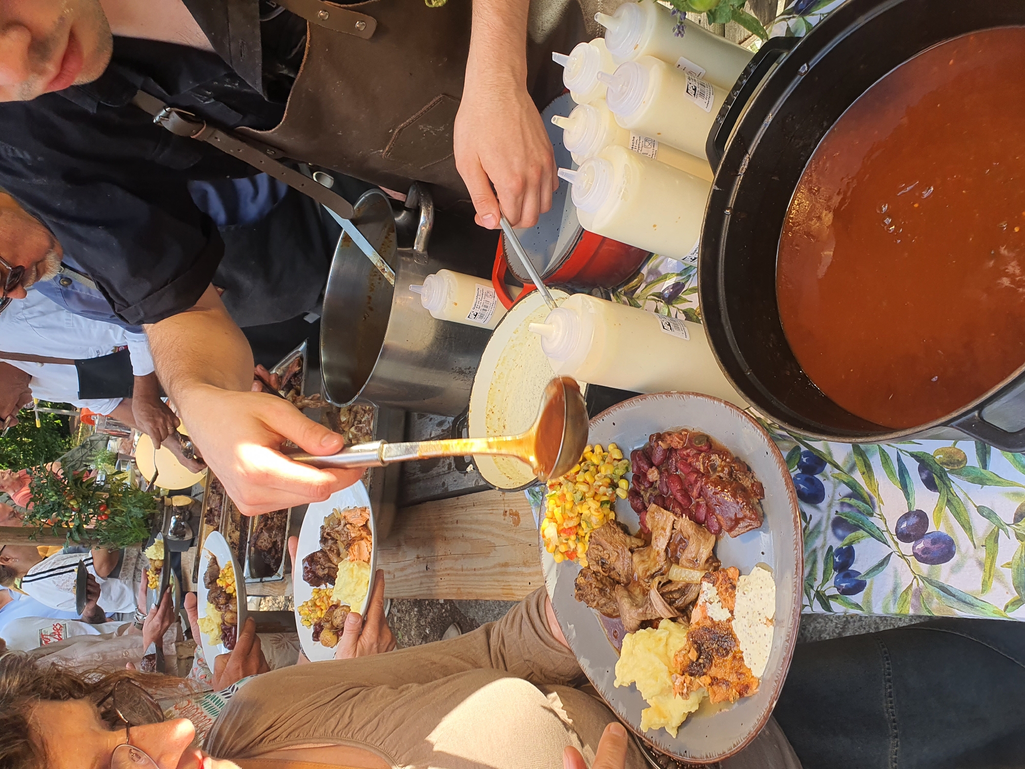 The image depicts an outdoor buffet or catered event, possibly in Falkensee, Germany, given the prevalence of such gatherings in the region. A woman in the foreground, wearing a light brown top and glasses, is holding a plate of food. The plate contains mashed potatoes, red beans, corn and bell pepper salad, and what appears to be roasted meat. To the right, a person wearing a dark apron is serving food with a ladle from a pot. Several squeeze bottles containing a white sauce are lined up on the table. In the background, other people are visible, some holding plates and engaging in conversation. The setting appears to be rustic, with wooden tables and a natural, outdoor environment. The overall atmosphere suggests a communal dining experience.