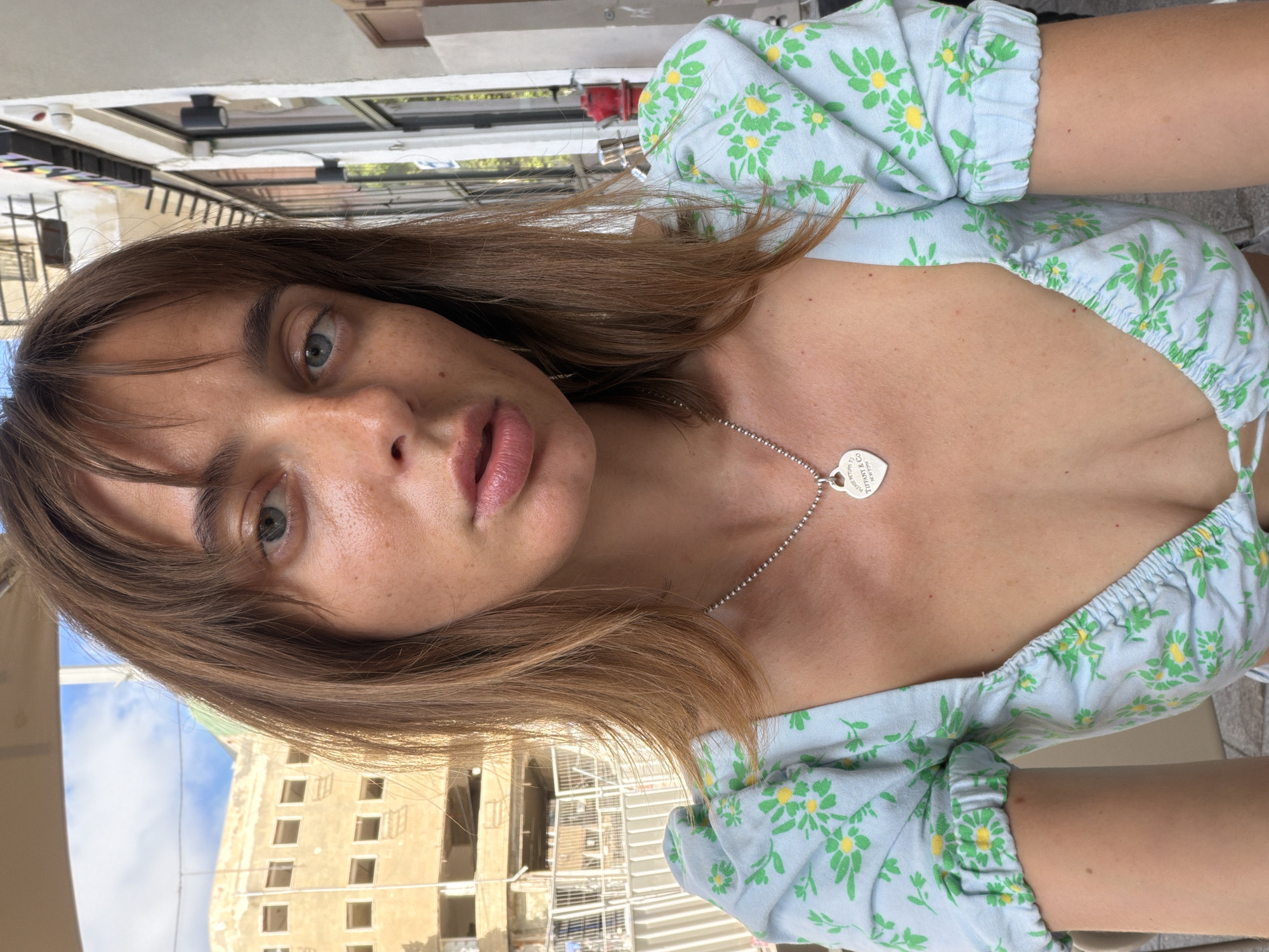 A young woman with shoulder-length, light brown hair and bangs is photographed from the chest up. She is wearing a light blue top with a green floral pattern. A silver heart-shaped pendant necklace with the inscription "Please Return to Tiffany & Co. New York" is visible. The background appears to be an outdoor setting in Tel Aviv, Israel, with a building under construction on the left and a storefront on the right. The lighting suggests it is daytime, possibly late afternoon given the angle of the sun.