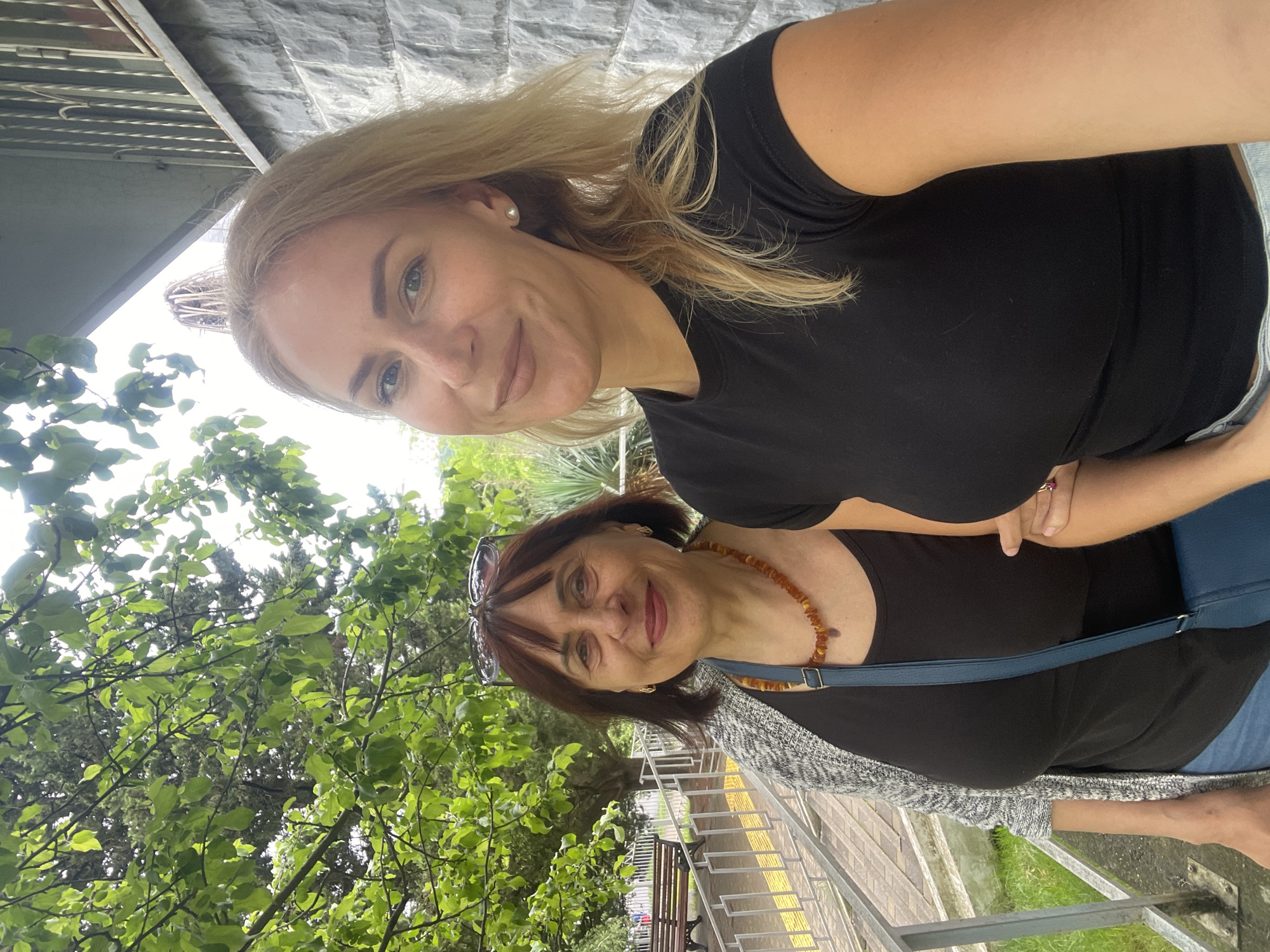 Two women are posing for a selfie. The blonde woman in the top of the frame wears a black shirt. The woman below has shoulder-length brown hair and wears sunglasses on top of her head, a black shirt, an amber necklace, and a gray cardigan. They are in Sochi, Russia, next to a metal railing and a walkway. In the background are trees and a gray stone wall.