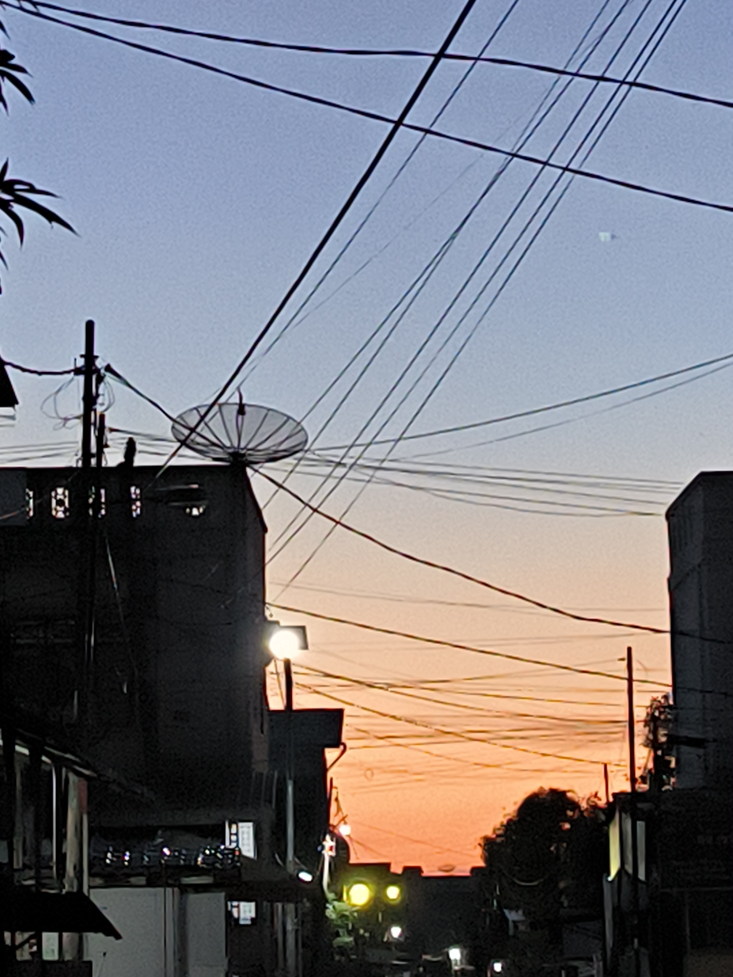 The photograph captures a street scene in Lhokseumawe, Indonesia, at either dawn or dusk, as indicated by the gradient sky from blue at the top to orange near the horizon. The cityscape is dominated by a dense network of overhead electrical and telephone wires that crisscross the view. In the foreground, several buildings, mainly in silhouette, line the street. One building features a satellite dish mounted on its roof. Streetlights are present and illuminated. A small tree is visible towards the center of the frame, situated along the street. No people are discernible in the photo.