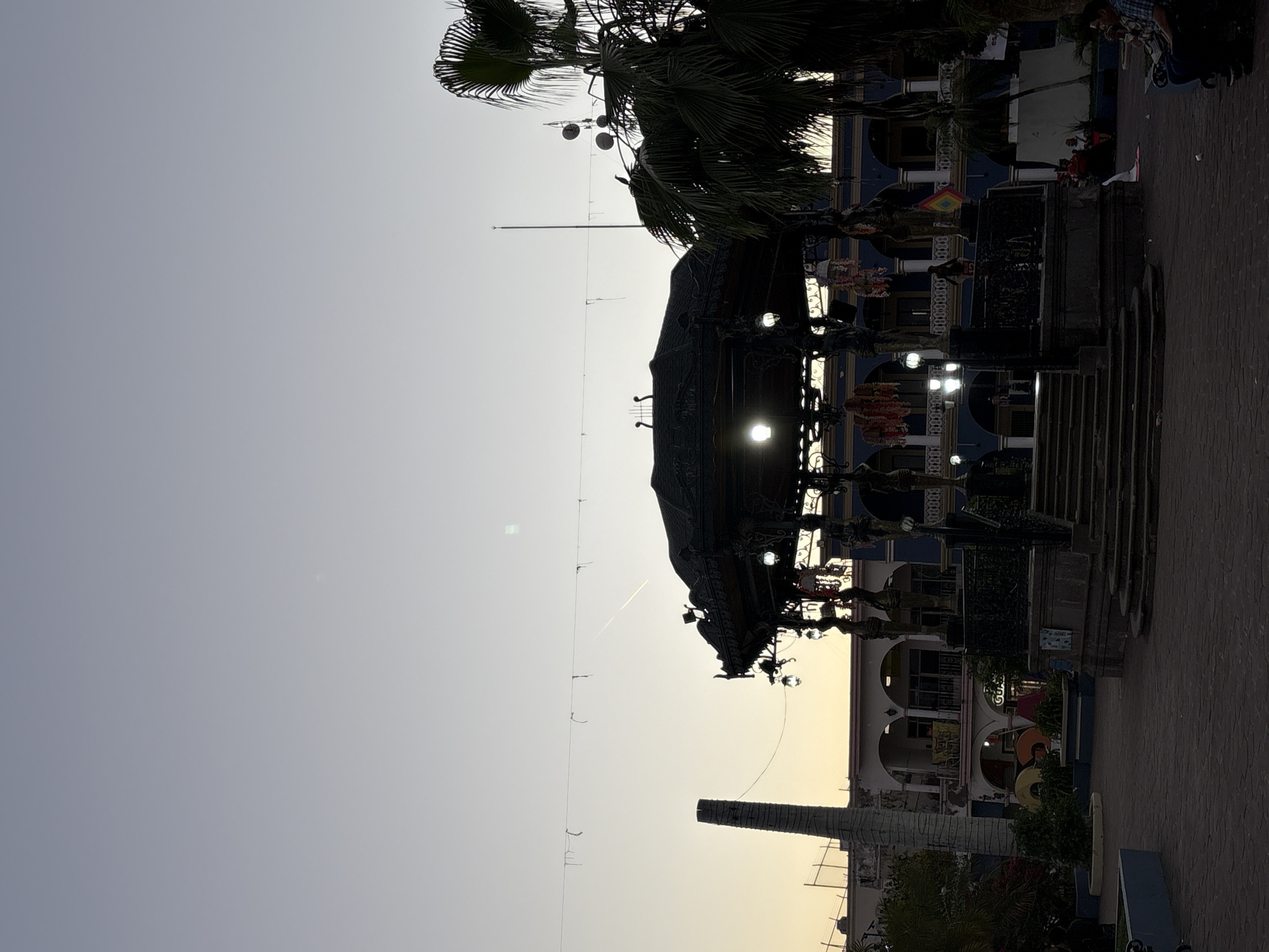 The image shows a plaza in Santiago Ixcuintla, Mexico, at dusk. Dominating the view is an ornate, wrought-iron gazebo or kiosk, with illuminated interior. Adjacent to the gazebo is a flight of stairs leading toward a building with arched windows and a blue and white facade. Palm trees are visible above the gazebo. A tall brick chimney is present behind the structure, silhouetted against the sky. Decorative lights are strung across the plaza. A small group of people is visible to the upper right. The paving appears to be brick or stone. The sky is a gradient of gray to yellow. A contrail is visible in the sky.
