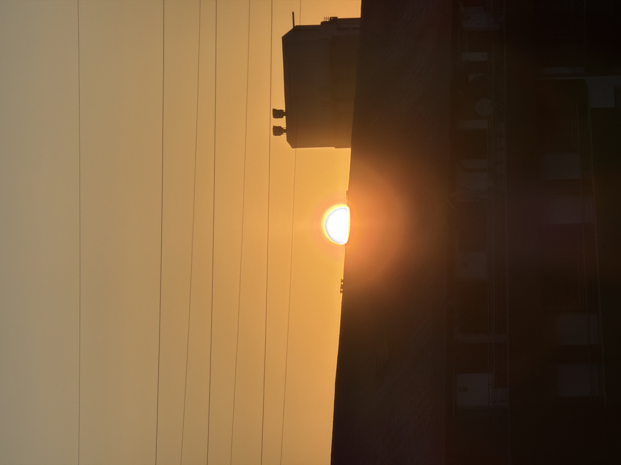 The image shows a sunset over a residential building in Esch-sur-Alzette, Luxembourg. The sun, partially obscured by a building in the foreground and a taller structure in the background, casts a warm orange glow across the sky. Several horizontal power lines stretch across the frame. The building in the foreground appears to be a multi-story apartment complex with balconies. The structure in the background has a distinct, boxy shape with two prominent vertical elements on its roof. The overall scene is silhouetted against the vibrant sunset, creating a dramatic and atmospheric perspective. There are no people visible in the image.