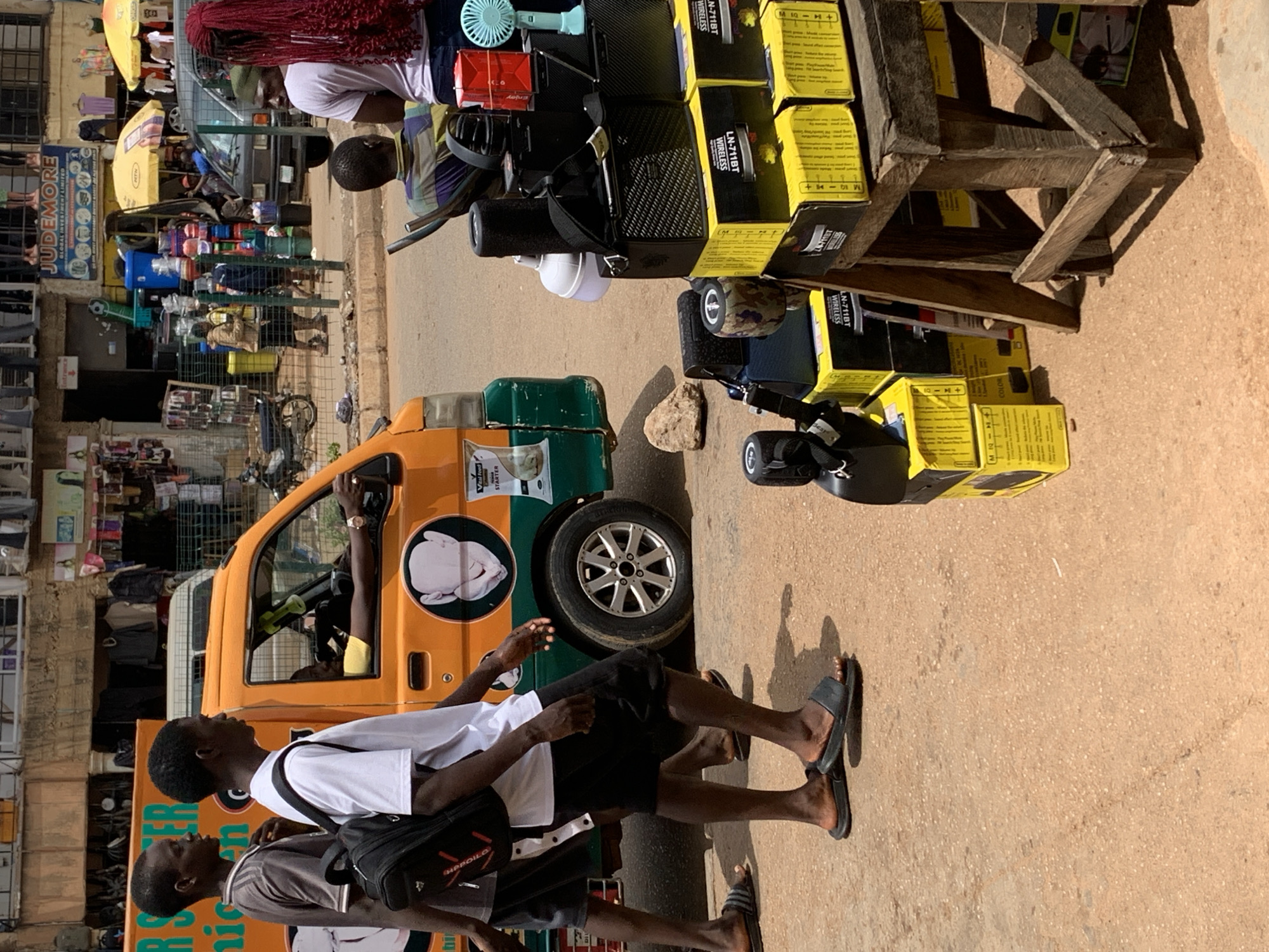 In Bukuru, Nigeria, two young men are walking on a dusty road beside an orange and green commercial vehicle. The man on the left wears a grey t-shirt and dark shorts, carrying a black backpack. The man on the right is in a white t-shirt and black shorts, also with a black backpack. Inside the vehicle, a man wearing a yellow shirt is visible through the driver's side window, appearing to be in control of the vehicle. The street is lined with buildings and shops, with various goods displayed, including electronics and household items. In the foreground, a makeshift wooden stand displays stacked yellow boxes, likely containing electronics, alongside other electronic devices like speakers and fans. A blue fan is also visible atop some of the boxes. In the background, several other individuals are present, some near a parked dark-colored vehicle. The overall scene depicts a busy urban environment with commercial activity.