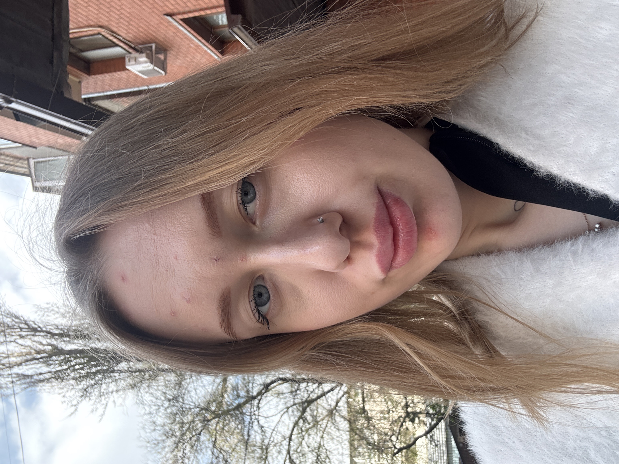 A close-up, portrait-style photograph shows a light-skinned young woman with light brown hair and blue eyes. She has a small silver nose piercing on her left nostril. She is wearing a fluffy white garment over a black top.
The background features a building with a red-tiled roof and what appears to be an air conditioning unit. Bare tree branches are also visible against a cloudy sky. The precise location within Rostov-on-Don, Russia is not discernable from the image.
