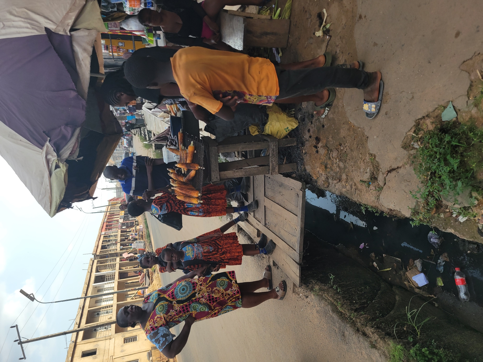 This media file depicts an outdoor scene in Egbagu, Nigeria, likely during daytime with bright sunlight casting shadows. The setting appears to be a street market adjacent to a drainage ditch filled with stagnant water and debris, suggesting an urban environment with potential sanitation challenges.
The primary subjects are several individuals, including adults and possibly children, engaged in commerce. One prominent activity involves the sale of corn on the cob. Several people are gathered around a vendor displaying an array of corn. Clothing is colorful and varied, with some individuals wearing vibrant, patterned dresses, while others are in simpler attire. The people seem to be interacting with each other, possibly as customers and vendors.
Objects visible include the corn being sold, a wooden stall or stand, a large umbrella providing shade, a yellow plastic bag, and a red plastic bottle in the drainage ditch. In the background, a multi-story building with balconies and windows is visible, indicating a built-up area. A street lamp with a curved pole stands prominently on the side of the road.
Notable details include the apparent bustling nature of the market despite the unsanitary condition of the nearby ditch. The expression on the faces of the people are not clearly discernible due to the image quality and orientation. There is no visible text in the image.
