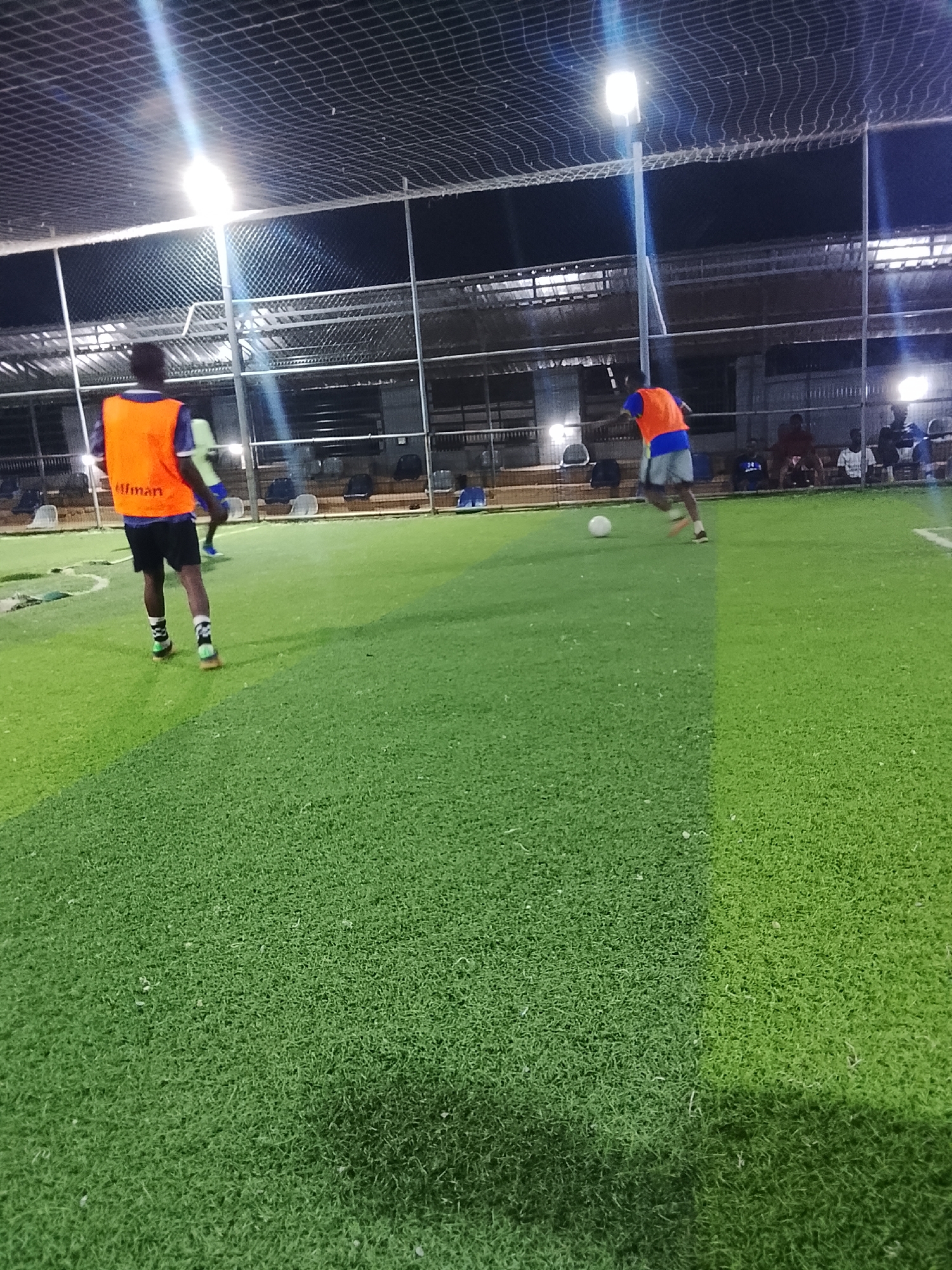 The image shows an evening indoor soccer game on a green artificial turf field, likely within the city of Akwanga, Nigeria, though there's no explicit visual confirmation of the location. The field is enclosed by netting and metal support beams, and stadium-style lighting illuminates the scene. Two players wearing orange vests are visible; one is dribbling the soccer ball, while the other is positioned further back. Several people are seated on chairs along the sidelines, appearing to watch the game.