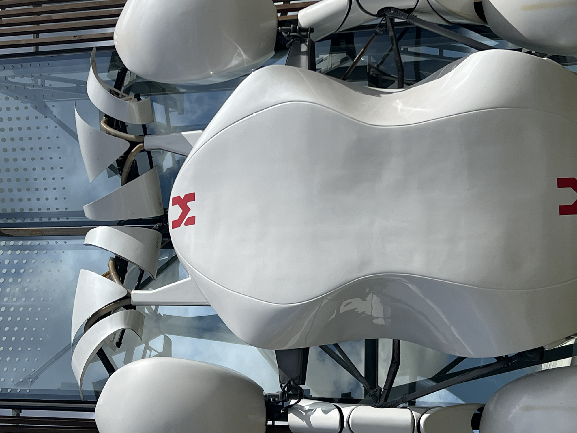 A white, abstract, sculpture-like structure is prominently featured in the center of the image. The structure has a rounded, bulbous main body with a red 'M' logo on its upper surface. It is supported by a visible metallic frame. To the left and right of the central body are large, white, spherical elements. The background consists of a modern building with a glass facade, reflecting a cloudy sky. The sculpture appears to be an outdoor installation in an urban setting. It is located in Veliky Novgorod, Russia.
