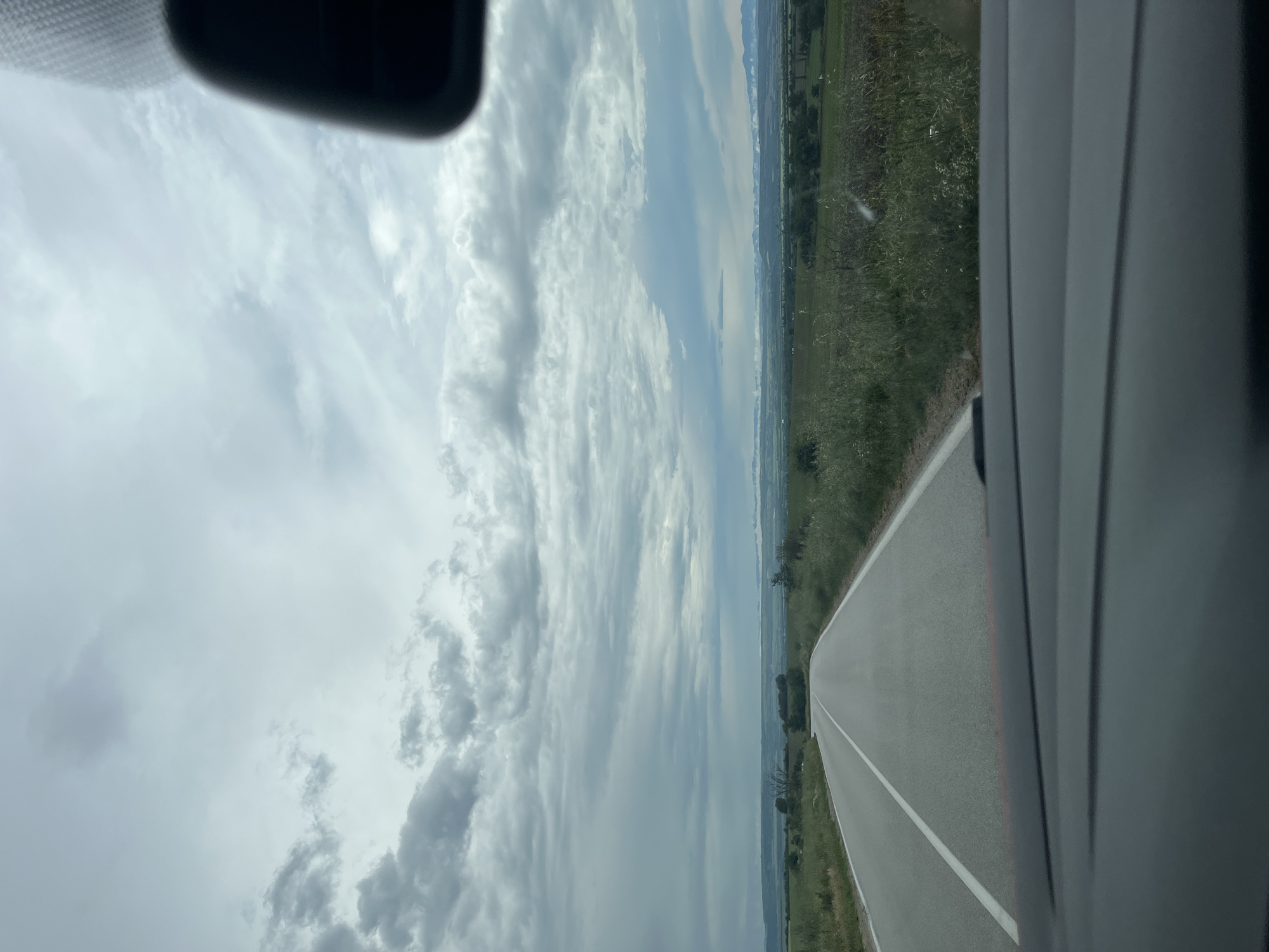 The image captures a rural landscape seen from inside a moving vehicle, likely looking out the front passenger side window. The perspective is oriented horizontally.
The upper portion of the frame is dominated by a vast, overcast sky filled with numerous layered clouds, ranging in color from bright white to various shades of gray, suggesting a heavily cloudy but dry day.
Below the expansive sky, an asphalt road stretches into the distance, appearing to curve slightly to the right. It features a clear white solid line down its center and a faint red or orange line near its right edge, likely indicating a shoulder. Lush green grassy verges line both sides of the road, beyond which lie wide, cultivated fields. In the far distance, a subtle line of low hills or mountains is visible against the horizon, partially obscured by atmospheric haze.
Portions of the car's interior are visible in the foreground. A dark, blurred shape, possibly part of a rearview mirror or dashboard, intrudes from the top left corner, while a dark, vertically textured element, likely the car's A-pillar or window frame, borders the right side of the image.
The scene depicts a peaceful journey through an agricultural region, consistent with the provided context of Resen, Bulgaria. The time of day is clearly daylight, under a heavily clouded sky. No people are visible, nor is there any discernible text within the image.