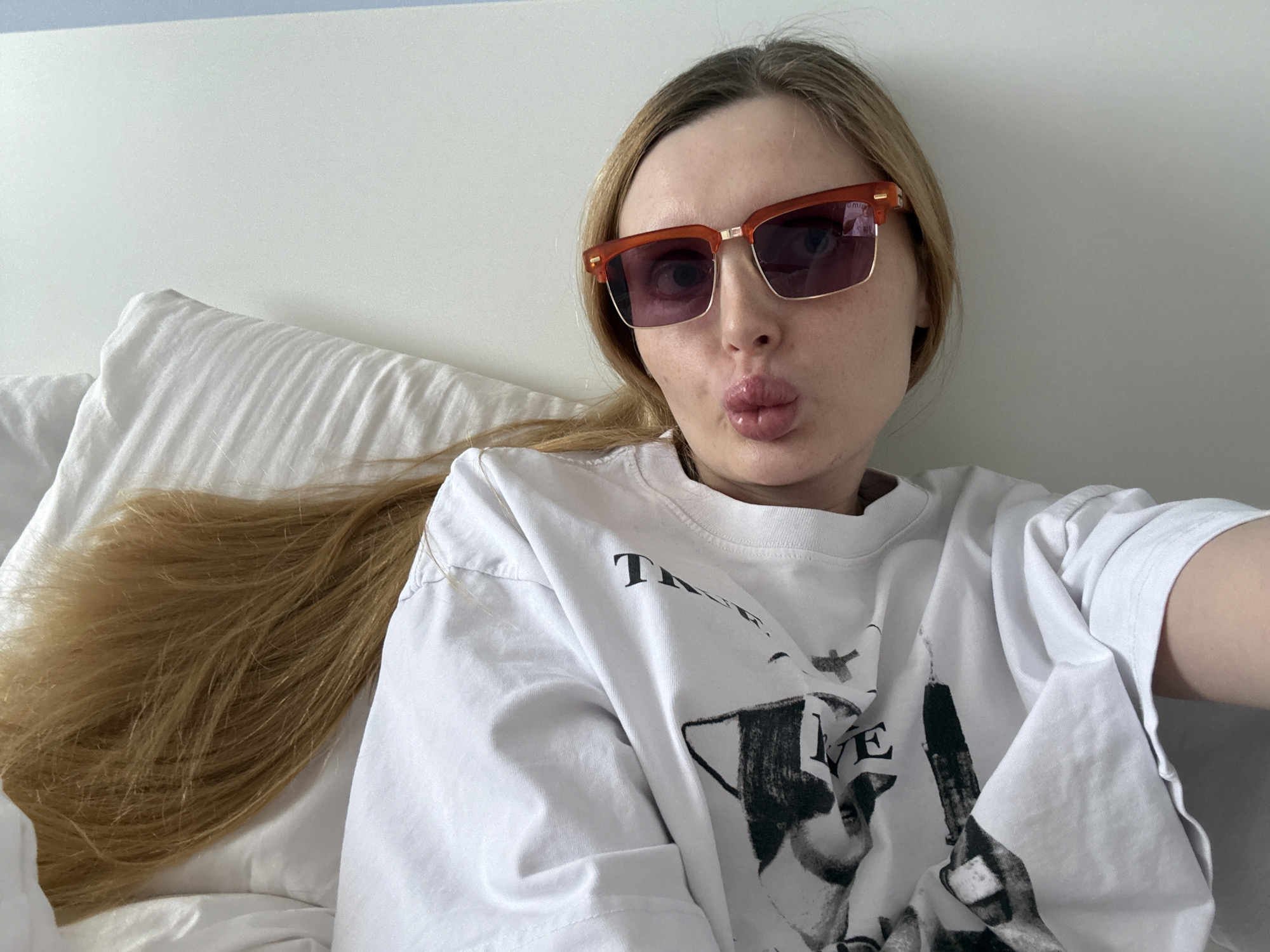 A young woman with blonde hair is lying on a white bed, wearing red-rimmed sunglasses and a white t-shirt with a black graphic print. She is making a kissing face towards the camera. Her hair is long and spread out on the pillow beside her. The background is a plain white wall and a white pillow.