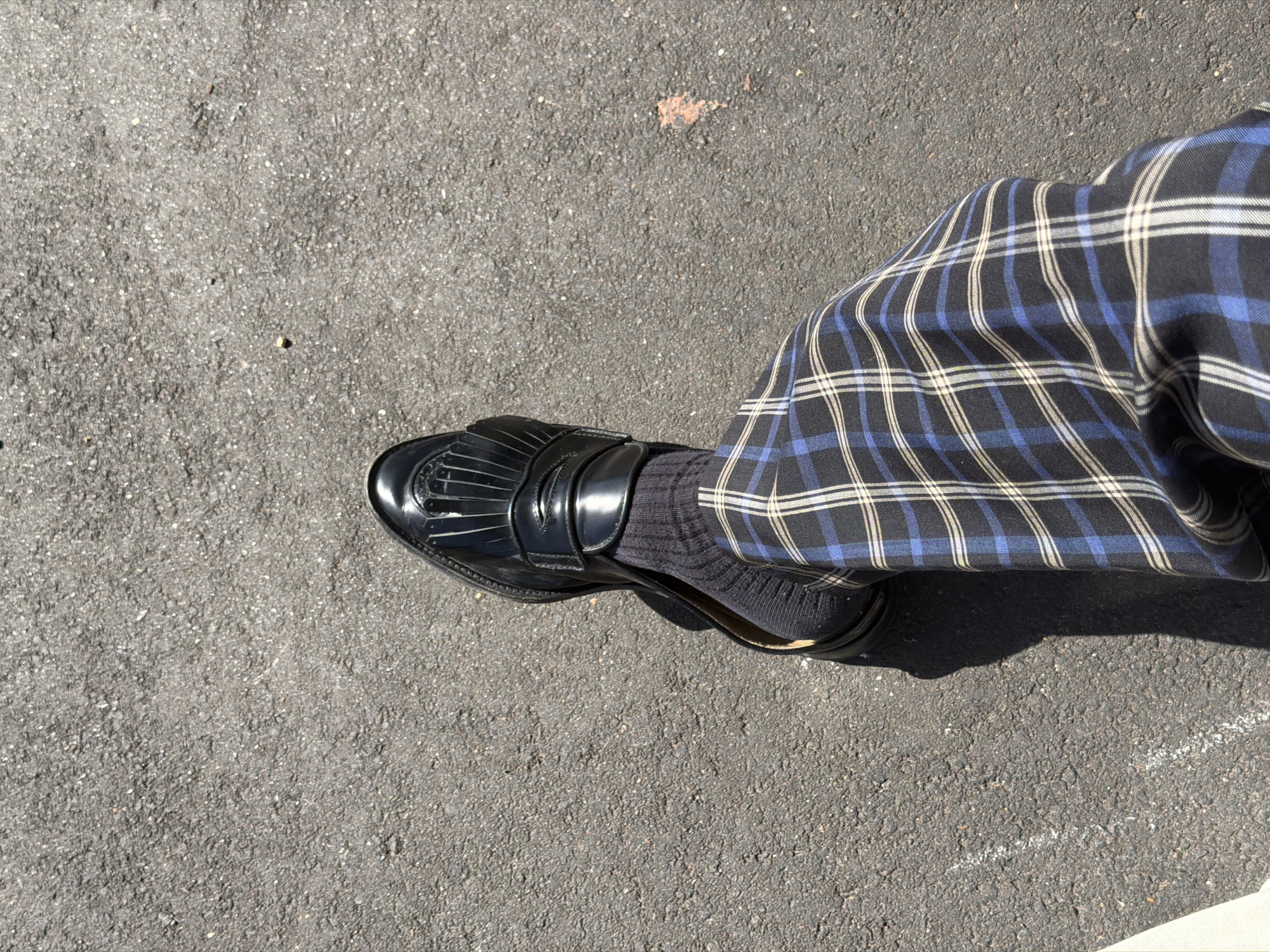 A high-angle, close-up shot captures a person's lower leg and foot resting on a dark grey asphalt surface. The individual is wearing a glossy black loafer shoe, detailed with a fringed or tasseled front and a strap across the instep. A dark, possibly ribbed, sock is visible between the shoe and the trouser leg. The trousers feature a bold plaid pattern in black, royal blue, and thin white or light cream lines.
The ground is a textured asphalt or tarmac, appearing dark grey with embedded lighter aggregate. Faint, irregular white lines, possibly chalk or faded paint, are visible on the asphalt in various spots. Bright sunlight illuminates the scene, casting a distinct, sharp shadow from the leg and shoe onto the ground, indicating a clear, sunny day outdoors.
The scene depicts a static moment, likely the person standing or taking a step. No specific activity or event is explicitly shown. The image is devoid of any visible text or discernible emotional expressions. The setting is an outdoor area, such as a path or street, consistent with the provided location context of the Folkestone and Hythe District, United Kingdom.