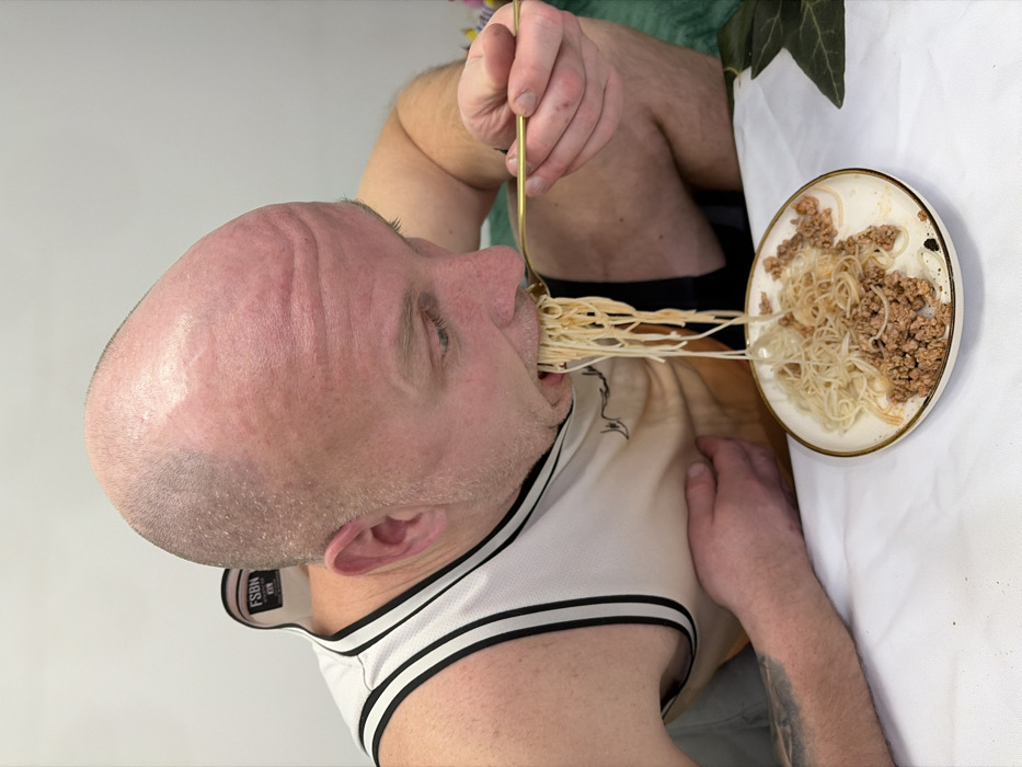 A bald man in a basketball jersey eats spaghetti with a gold fork. He's seated at a table covered with a white cloth. The plate of spaghetti also contains meat sauce. A small green leafy plant sits on the table, obscuring the background. The man's head is tilted back as he consumes the pasta.