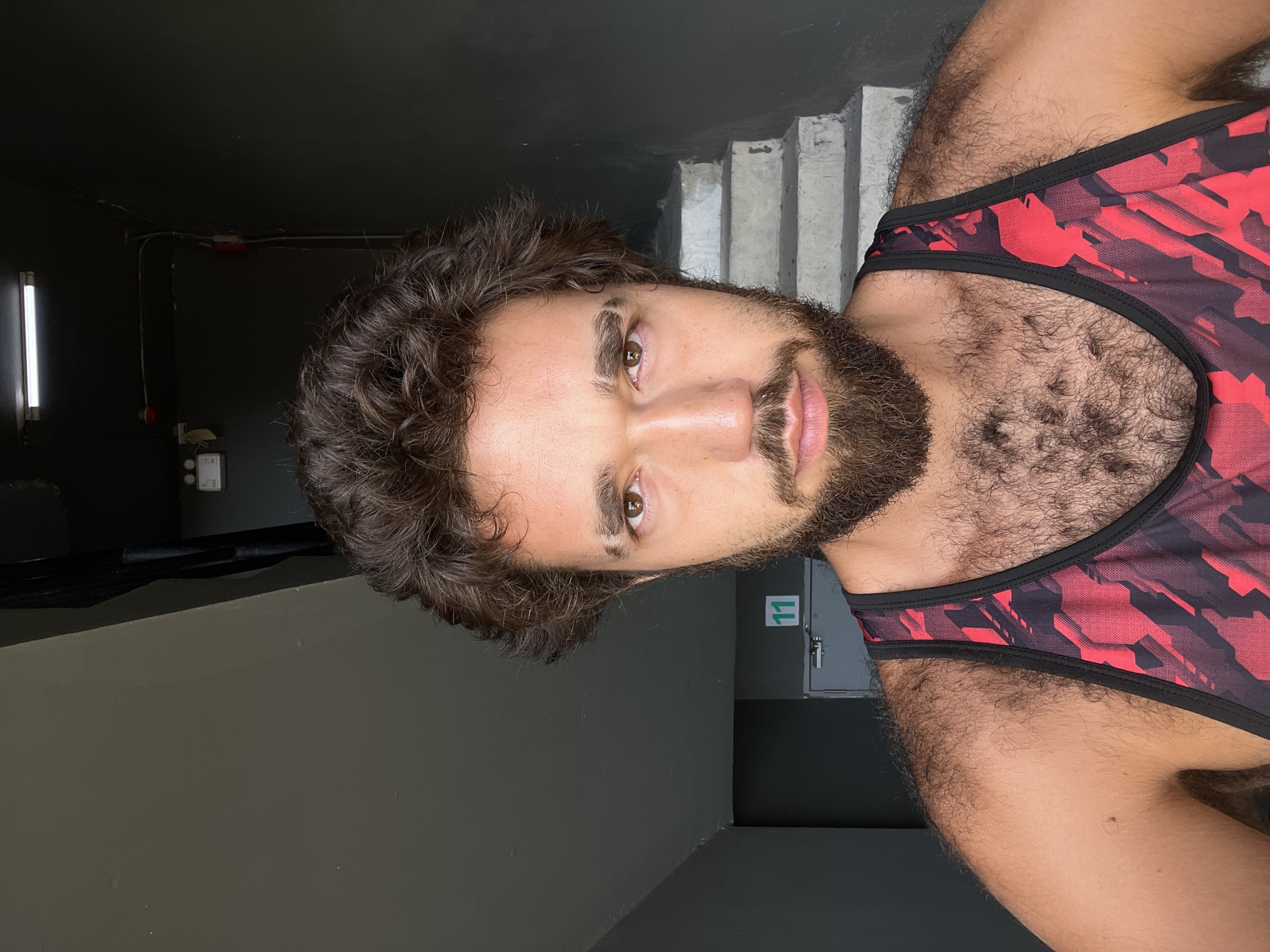 A close-up, rotated portrait shows a young man with dark, curly hair and a beard. He is wearing a red and black camouflage-patterned athletic tank top. The man has light brown eyes and appears to be looking directly at the camera. The background is dimly lit, with a dark wall and a set of concrete stairs visible. A white light fixture is mounted on the left side of the frame. Further in the background, a gray door with a green sign displaying the number "11" is visible. The scene suggests an indoor, possibly utilitarian or athletic environment, with elements that could be found in a gym or a stairwell. The lighting is somewhat harsh, creating shadows. There are no obvious indicators of time of day or weather conditions, but the overall mood is direct and unposed. The context places this scene in Bangkok, Thailand.