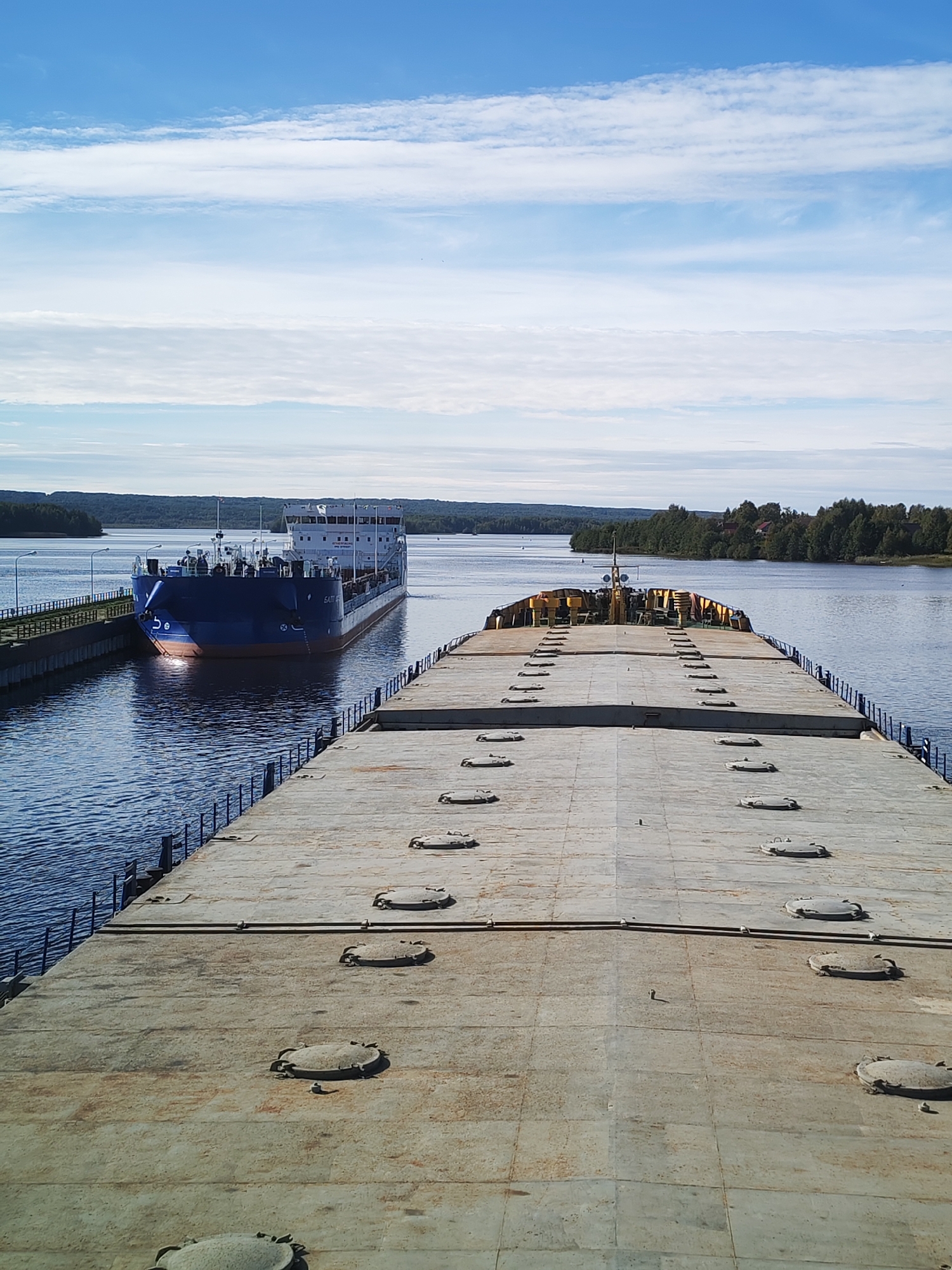 This image shows a view from the deck of a large, flat barge on a calm, blue river, likely in Анхимово, Russia. The barge's weathered, concrete deck occupies the foreground, with numerous circular hatches visible. In the middle ground, a blue cargo ship is entering or exiting a canal lock, with its bow facing towards the viewer. The ship has white superstructures and appears to be a tanker or a similar vessel. The canal walls and railings are visible on the left side of the image. The background features a tree-lined shore and a vast, partly cloudy blue sky. The overall scene depicts a quiet, sunny day on the waterway, with maritime traffic evident.