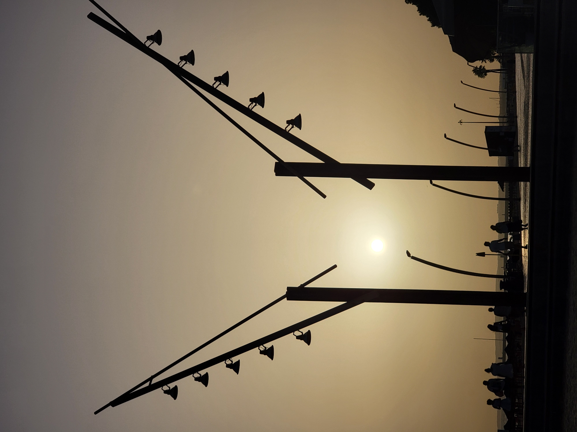 A silhouette of a coastal promenade in Quarteira, Portugal, at sunset. The sky is a pale gradient of beige and gray, with the bright, diffused sun near the horizon. Two large, V-shaped lampposts dominate the foreground, their dark metallic structures arching upwards and outwards, each adorned with multiple lamps. In the middle ground, a line of people, also in silhouette, stroll along the promenade, some standing and conversing, others walking. Further in the distance, more lampposts, some curved, are visible along the waterfront, with a hint of the sea to the left. A dark, amorphous mass, possibly a building or a hillside, forms the right edge of the frame. The overall impression is one of a tranquil evening scene at a popular seaside location.