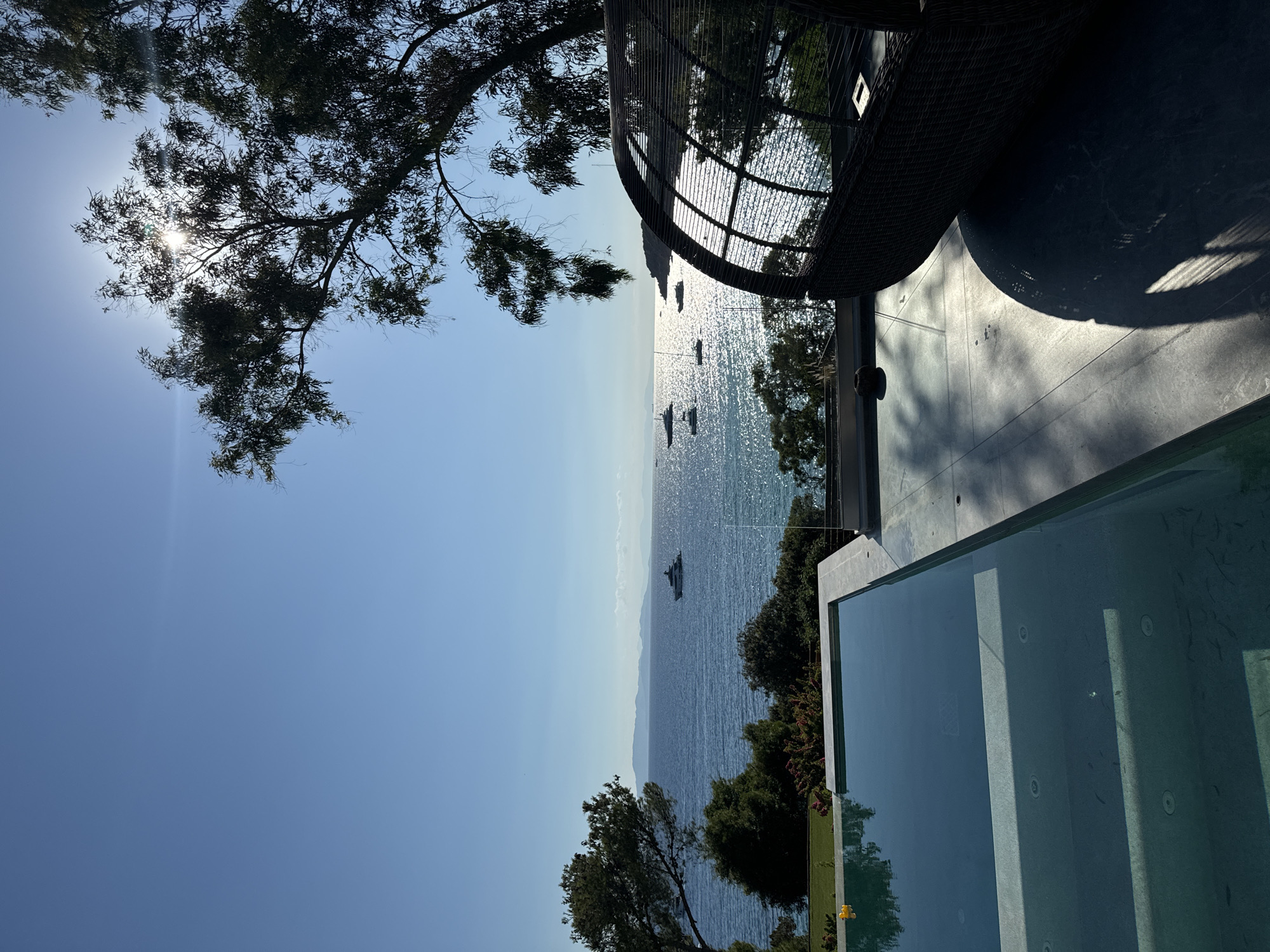 This image depicts a serene coastal view from a modern villa in Vouliagmeni Municipal Unit, Greece. In the foreground, a clean-lined infinity pool with clear, blue water reflects the sky and surrounding trees. The pool's edge is defined by light-colored stone. Adjacent to the pool, a contemporary outdoor lounge area features a large, dark woven pod-shaped chair casting a long shadow on the tiled patio.
The middle ground transitions to a lush green landscape with trees and bushes, leading to the sparkling blue sea. Several yachts and smaller boats are scattered across the water, some appearing as distant silhouettes against the bright, sun-drenched surface. The sun is high in the sky, partially obscured by the branches of a large tree in the upper right quadrant, creating a lens flare effect. In the background, a distant landmass with mountains is visible on the horizon under a clear, bright blue sky. The overall atmosphere is one of luxury, tranquility, and a Mediterranean summer day.