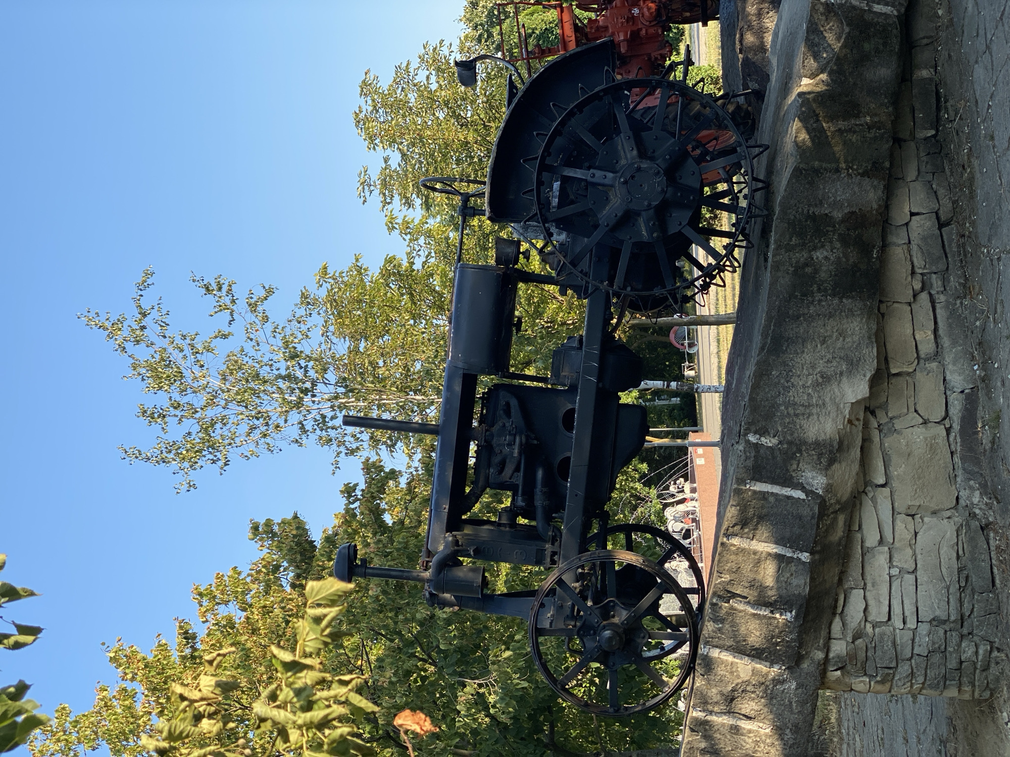 A black, vintage tractor is displayed outdoors, mounted on a rustic stone structure. The tractor, weathered and dark, is positioned at an angle, showcasing its large spoked wheels and engine components. Behind the tractor, lush green trees and a clear blue sky form the backdrop. To the right, a portion of a red tractor is visible. The scene appears to be in a public space, possibly a park or an outdoor exhibition, given the surrounding foliage and paving stones. The overall impression is one of historical preservation and display. This tractor is located in the Museum of the History of the City of Chisinau, Moldova.