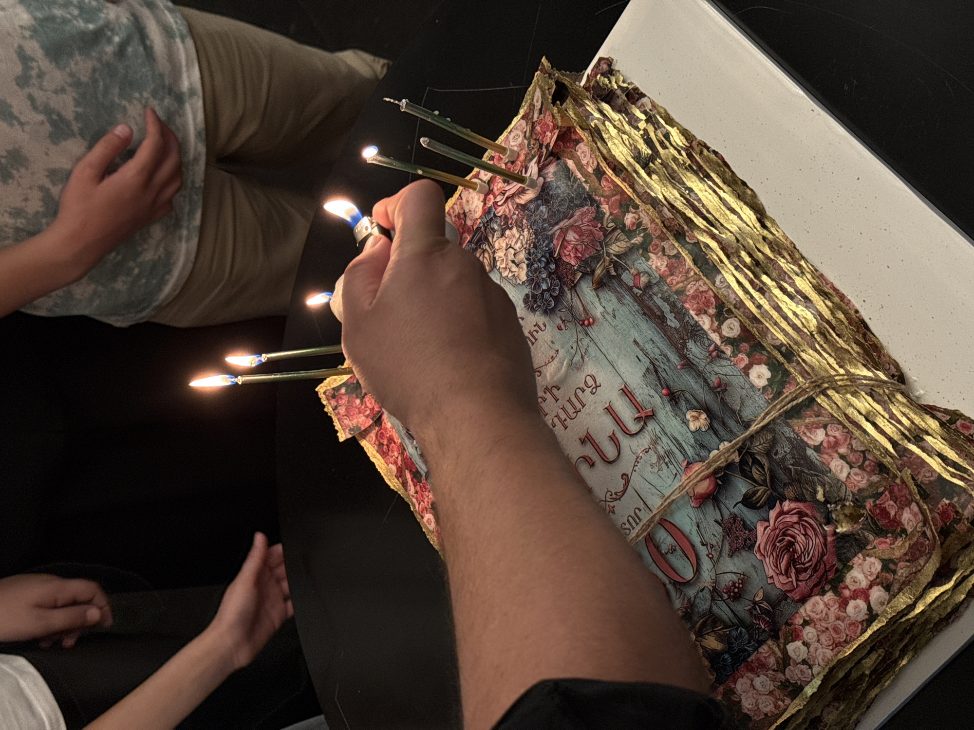 An indoor scene captures a close-up of a person's hand lighting a candle inserted into a highly decorative book or stack of papers. The hand, from the right foreground, holds a blue and silver lighter to ignite one of several thin, green candles. Three other candles are already lit, their small flames illuminating the immediate area.
The central object is a unique, ornate book or bound stack of papers, adorned with intricate floral patterns featuring pink and blue roses, berries, and leaves on a faded blue-green background. It appears to be tied with rustic twine, and the edges of its pages are elaborately gilded in gold. Visible Armenian script on the cover includes the letters "ԹԱՐՄ" (tarm, meaning "fresh"), "ՄԱՆ", "ՅԱՆ", and the number "0".
In the background on the left, another person is partially visible, wearing a light green camouflage-patterned t-shirt and tan pants, with their hand resting on their lap. A third person's hand and forearm can be seen in the lower-left corner of the frame, wearing a white shirt.
The setting suggests a dim environment, likely evening or night, with the candle flames serving as primary light sources. The activity is taking place on a dark, possibly black, surface, with a lighter-colored, possibly white, surface visible to the right of the book. This event is observed in Yerevan, Armenia.