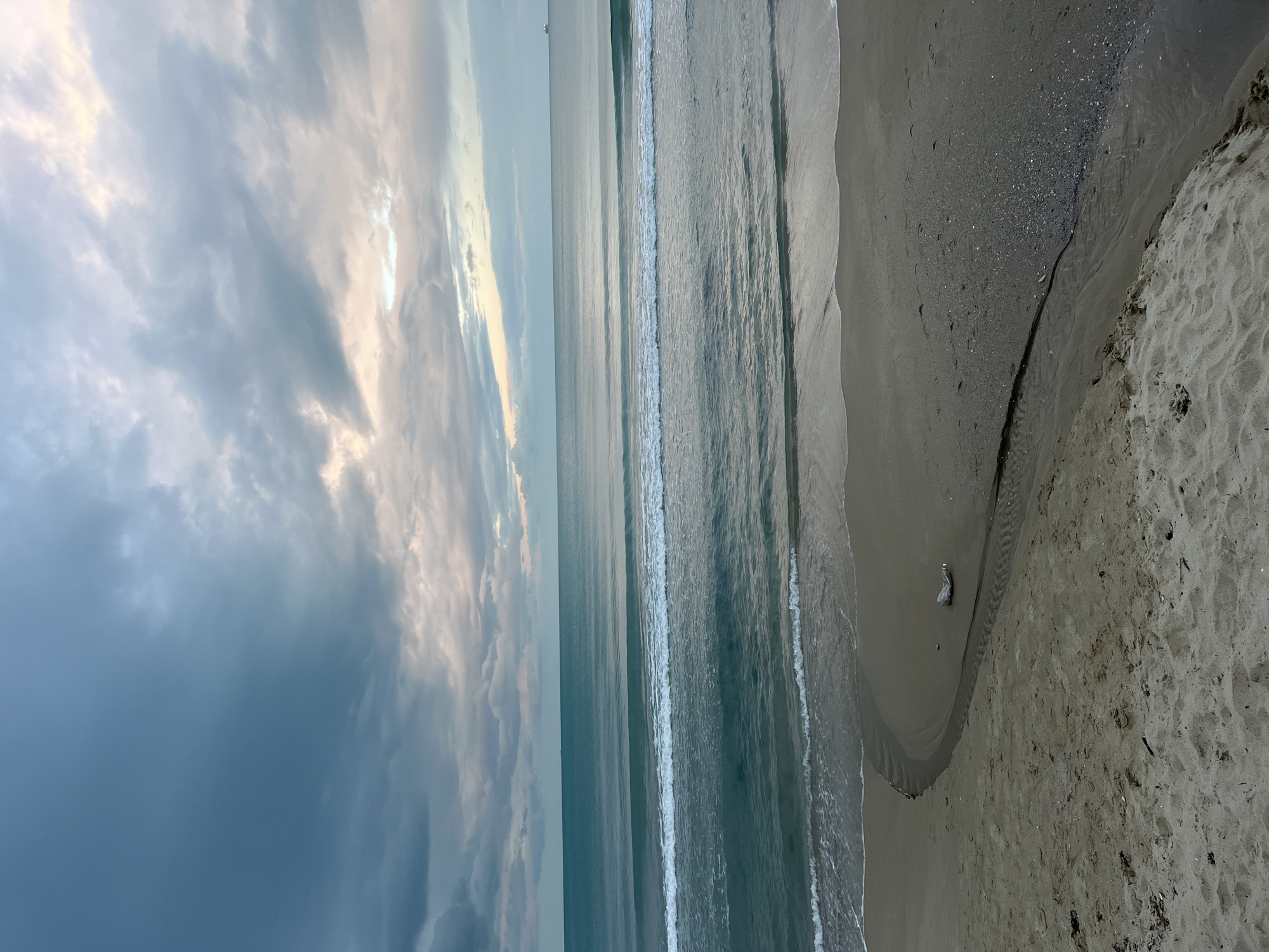 The image shows a beach scene in Varna, Bulgaria, at dusk or dawn. The sky is predominantly cloudy with patches of light breaking through. The sea is calm with gentle waves reaching the shore. The sand is wet near the water and becomes drier further inland. A small stream or runoff has carved a path through the sand towards the sea. A discarded plastic bottle is visible on the sand near the stream's mouth. There are no people visible in the image.