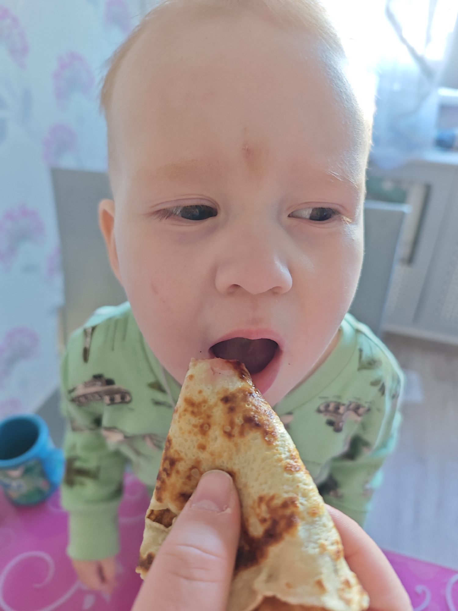 A close-up shot focuses on a young child eating a triangular pancake, which is being held by a hand. The child has fair skin, light red hair, and is wearing a green long-sleeved shirt with a tank print. The background features a blue and white floral wallpaper, with a gray chair and a blurred white radiator. The scene is indoors, with a pink table and a blue cup visible in the lower left corner. There is no verifiable evidence to confirm the location is in the city of Markovskii, Russia.