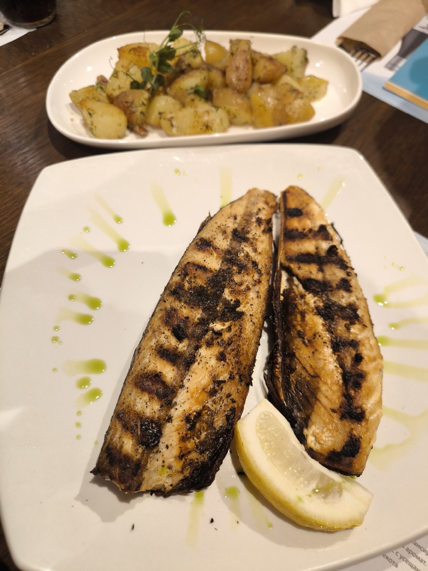 The image shows a plate of grilled fish with a lemon wedge and an accompanying side dish of diced potatoes with herbs. The fish, presented as two fillets, is heavily marked with grill lines. The potatoes appear golden and are garnished with sprigs of a green herb. The food is placed on a square white plate, with traces of green oil or sauce. In the background, a white oval plate holds the diced potatoes. The image was taken indoors, presumably in a restaurant. The location is Unknown, Unknown.
