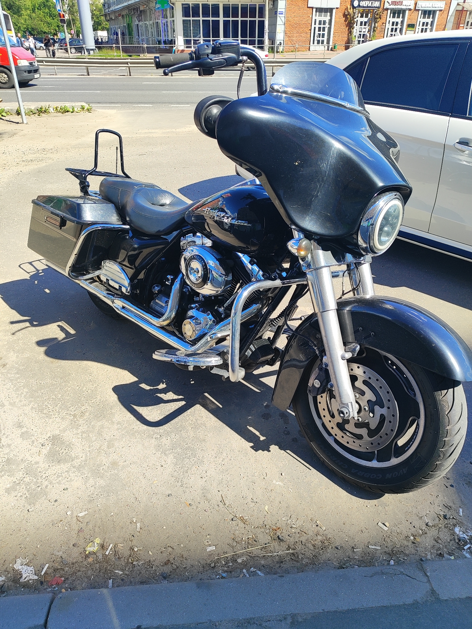 A black Harley-Davidson motorcycle is parked on a paved surface, casting a long shadow to the left. The motorcycle has a large black fairing, a comfortable-looking seat, and two saddlebags. Chrome accents are visible on the engine, exhaust pipes, and handlebars. A white car is parked behind the motorcycle. In the background, there is a street with traffic, including a red van. Buildings with storefronts line the street. The scene appears to be in an urban environment. The time of day is likely daytime, given the bright sunlight and shadows. The weather appears to be clear and sunny. The location is Shchyolkovo, Russia. There is some trash on the ground near the curb. Text is visible on the motorcycle's engine cover, appearing to say "Harley-Davidson". The tire of the motorcycle has text that reads "AVON COBRA CHROME".
